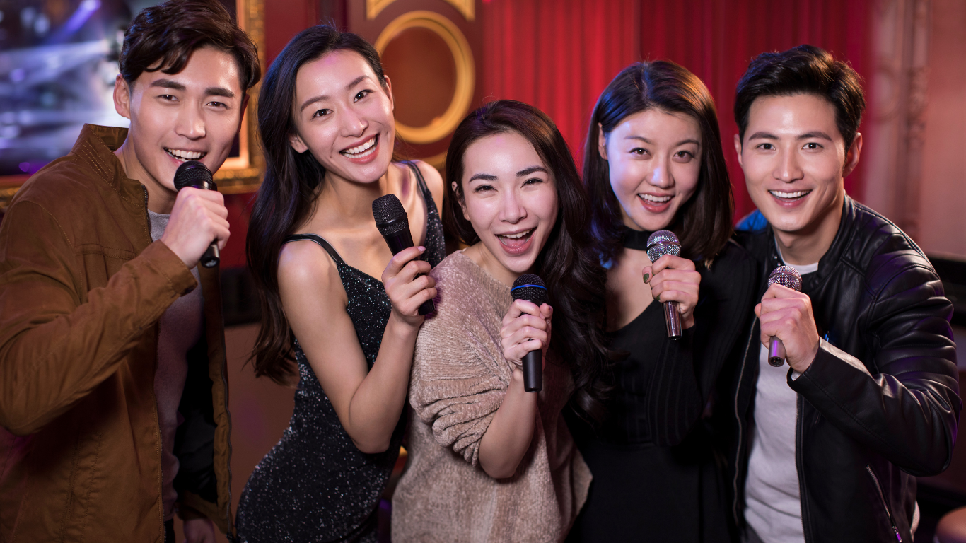 Group of five people holding microphones and smiling, appearing to sing together in a karaoke setting with a dimly lit, festive background.
