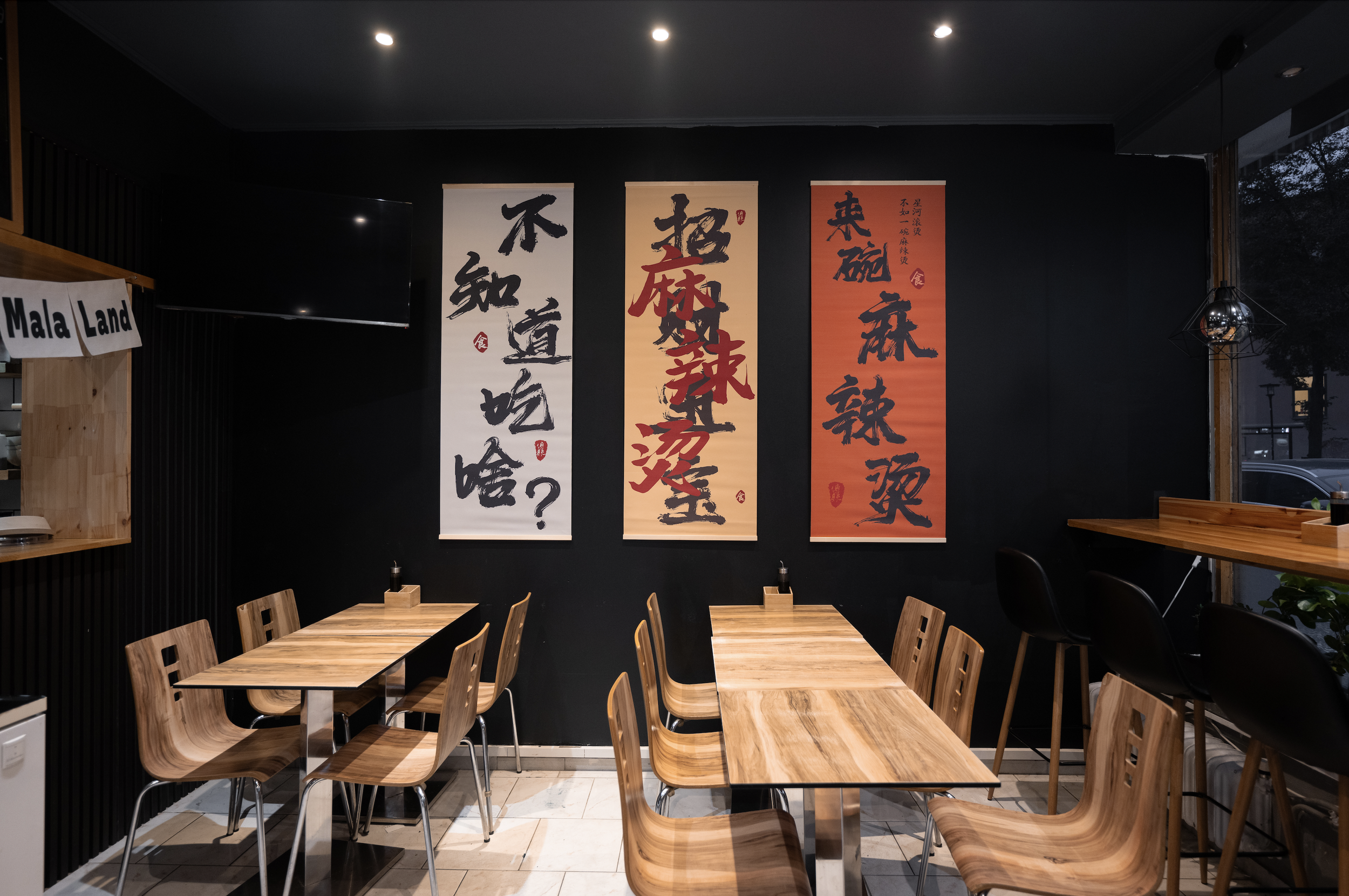 Interior of a restaurant with wooden tables and chairs, wall art with Chinese calligraphy, and a small sign reading 'Mala Land'.
