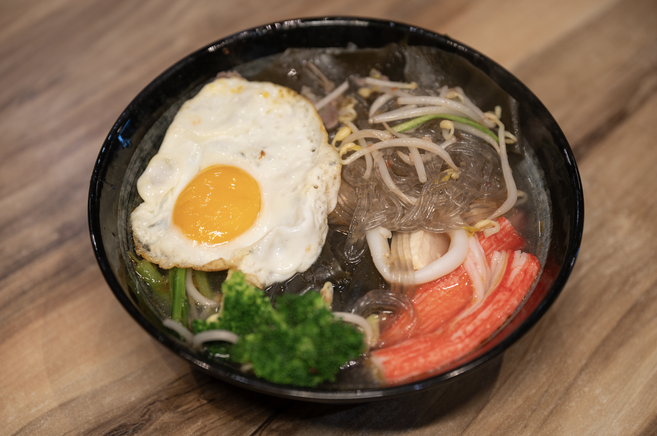 A bowl of Asian noodle soup with a fried egg, clear noodles, bean sprouts, imitation crab sticks, broccoli, and seafood pieces.