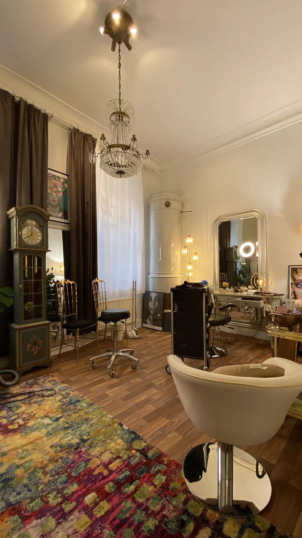 Interior of a stylish salon with a chandelier, ornate grandfather clock, colorful rug, mirrors, and salon chairs.