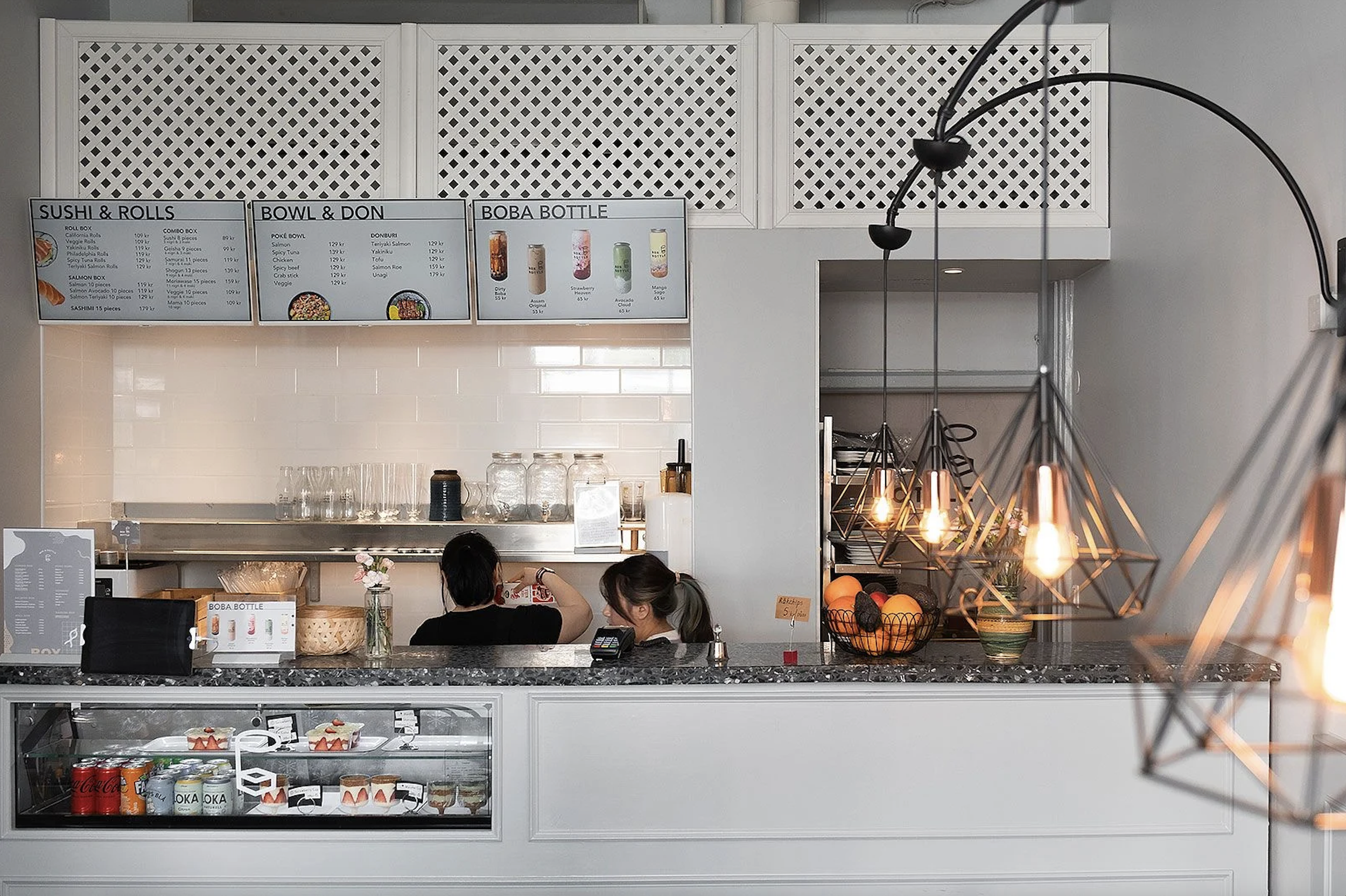 Interior of a modern cafe with menu boards for sushi, bowls, and boba bottles, counter with food display, and geometric pendant lights.