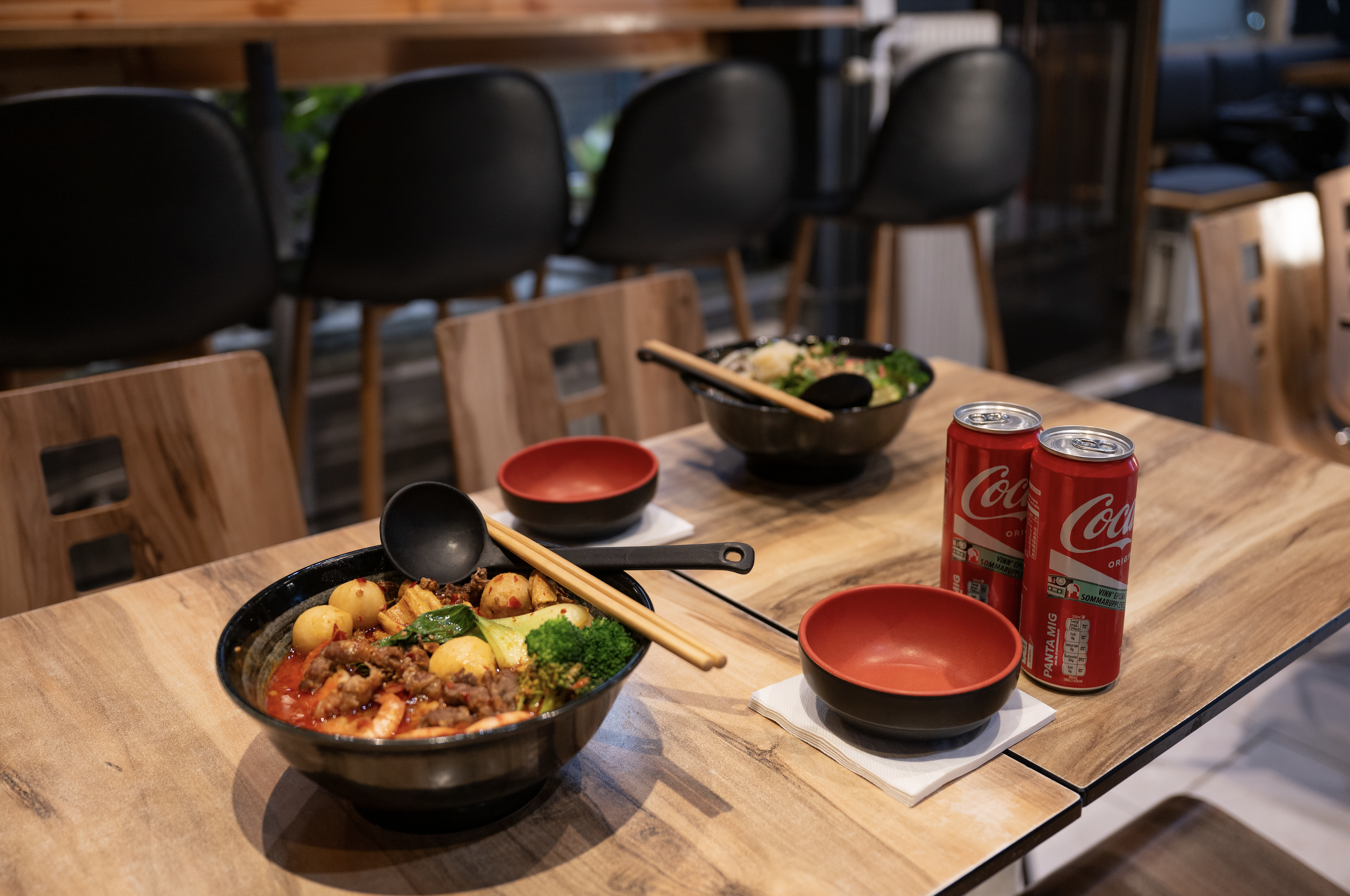 Two bowls of Asian-style noodles with vegetables and meat on a wooden table, accompanied by chopsticks and a black ladle, two red bowls, and two cans of Coca-Cola.
