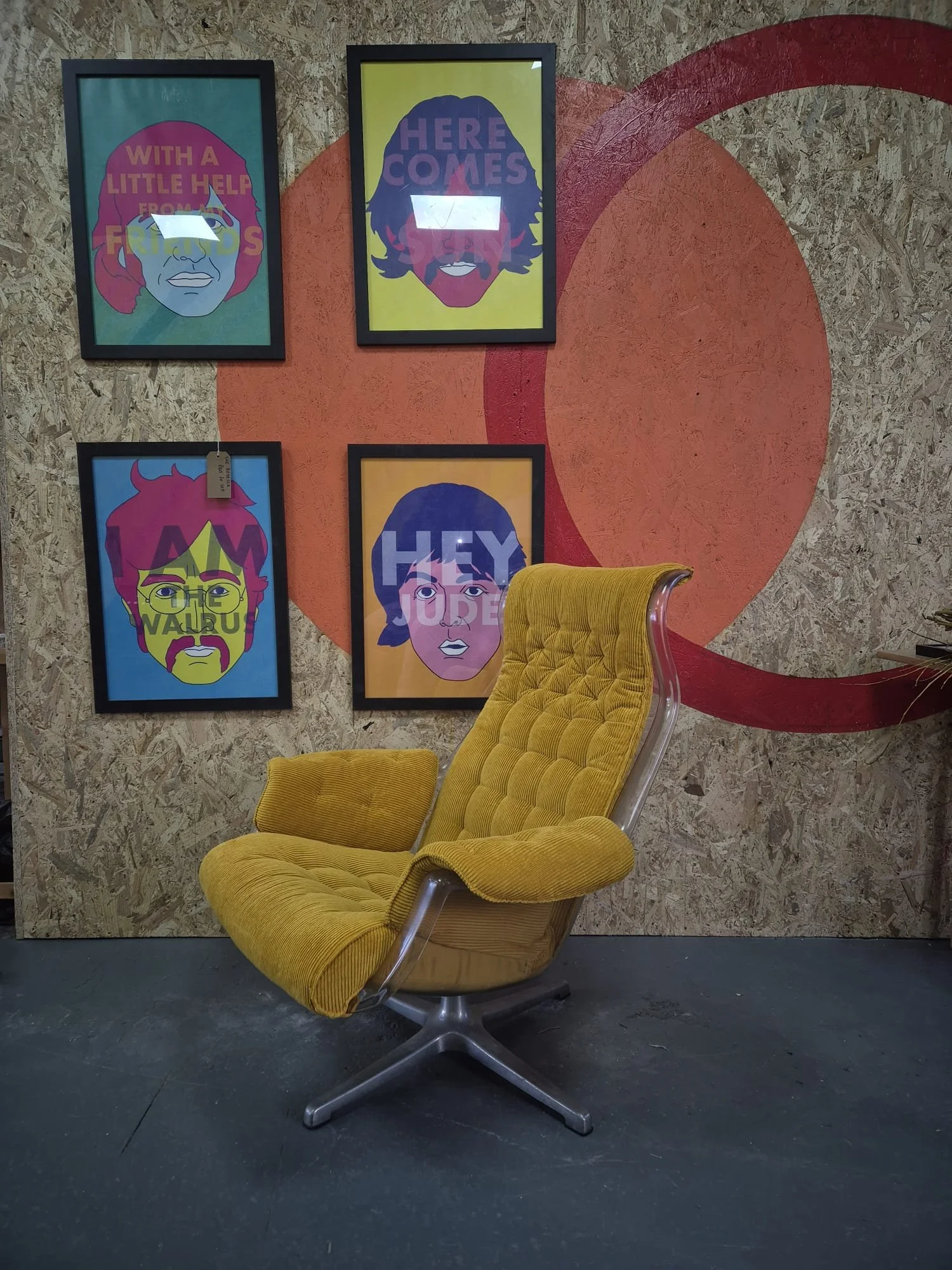 A yellow vintage Alf Svensson galaxy chair with a tufted backrest and armrests, positioned in front of a wall decorated with five framed colorful pop art portraits of cartoon faces with speech bubbles. based in an upholstery studio in Stockport.