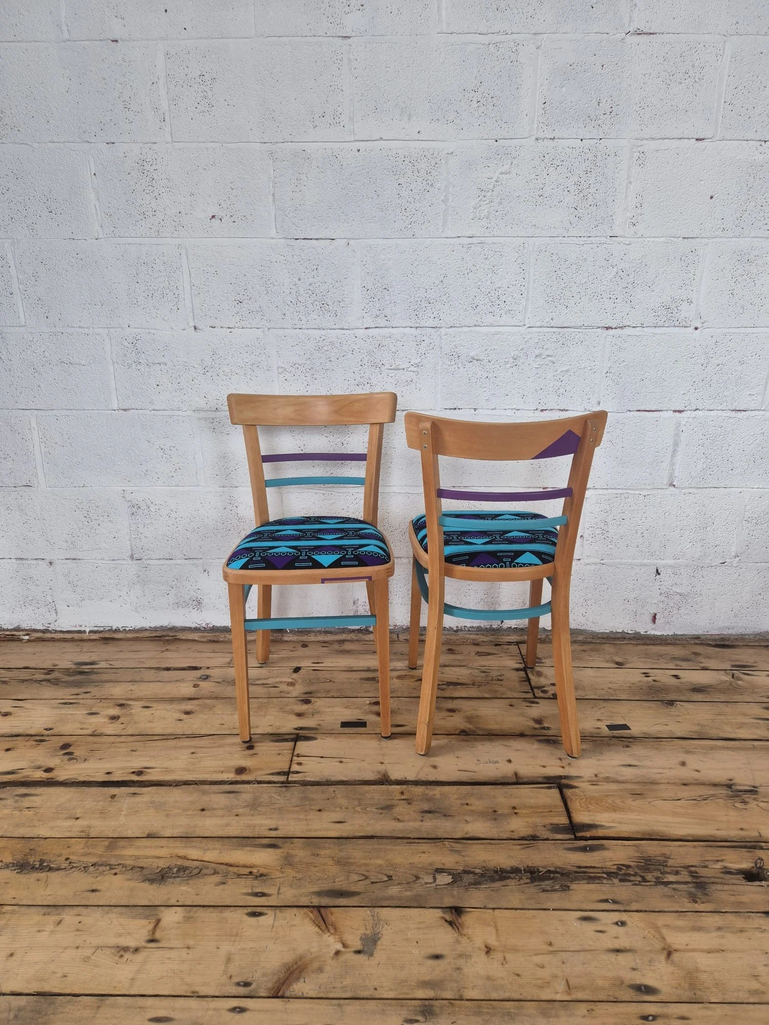 Two wooden chairs with colorful patterned cushions, placed on a wooden floor against a white brick wall.