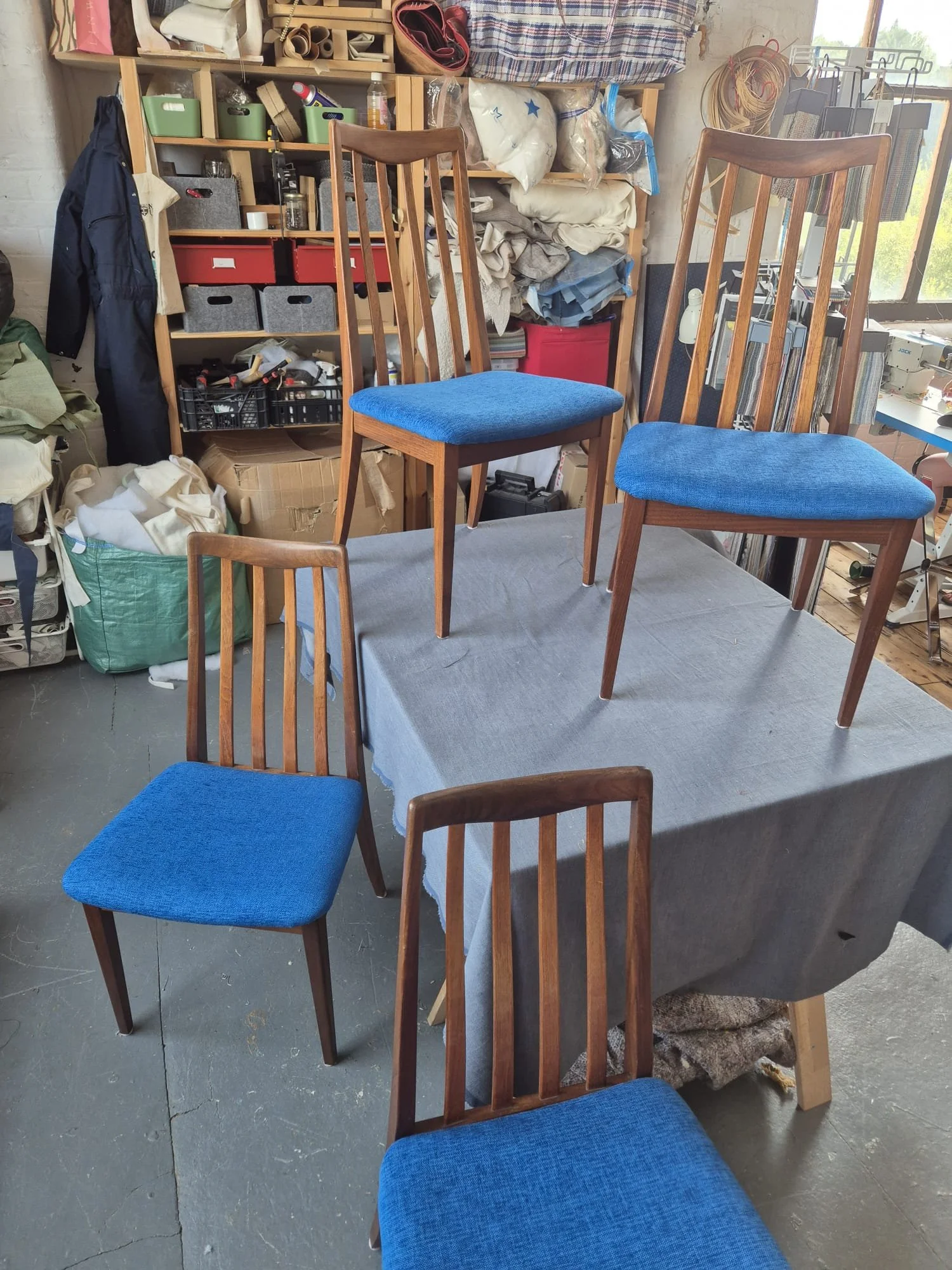 A set of four wooden chairs with blue cushioned seats arranged on and around a table covered with a gray cloth. The background shows a workshop with shelves, storage boxes, and various tools and materials.