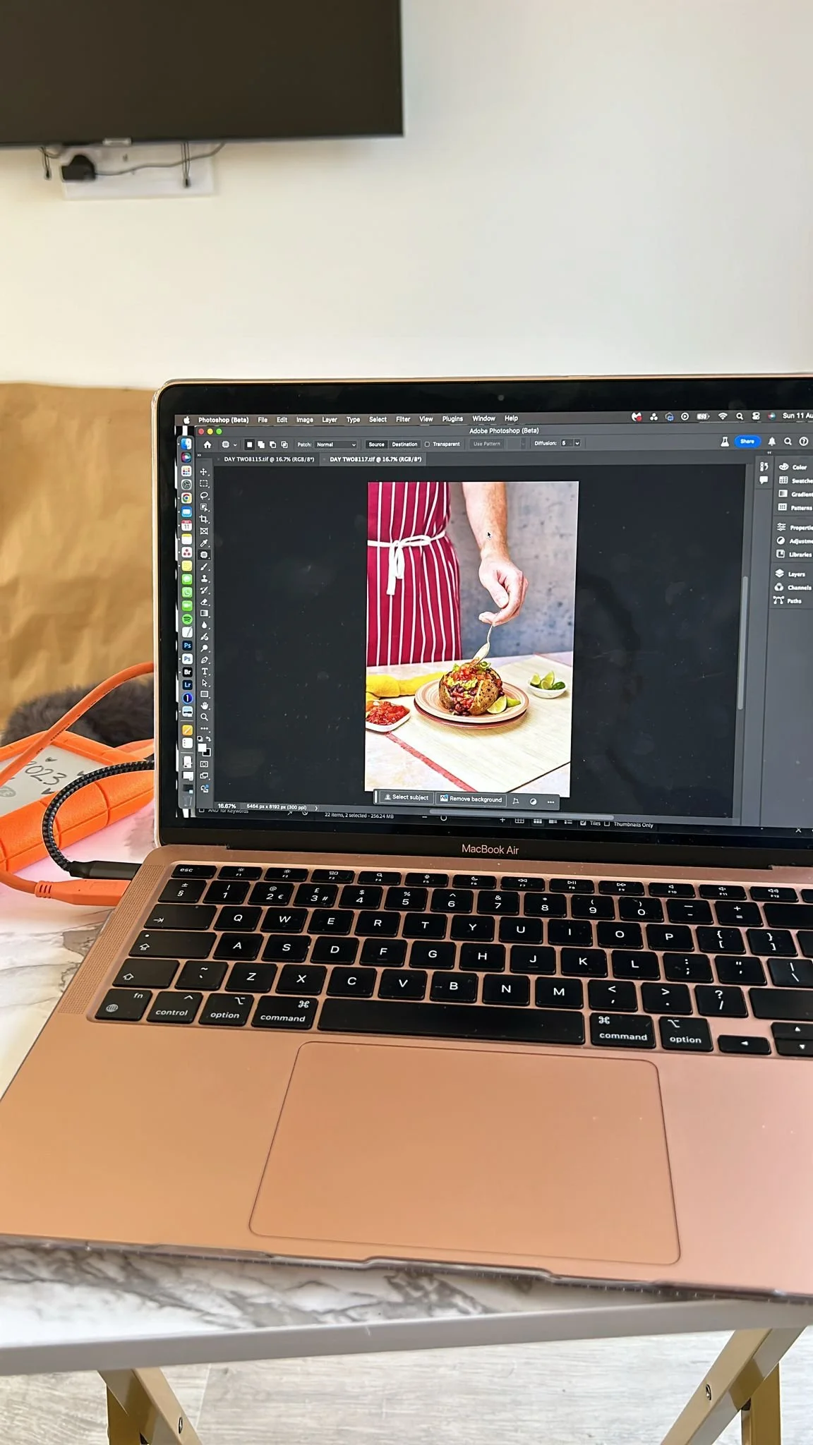 A MacBook Air laptop on a marble table displaying a photo of a person in a red apron holding a spoon over a plate of food, with a background showing a white wall and a black TV.
