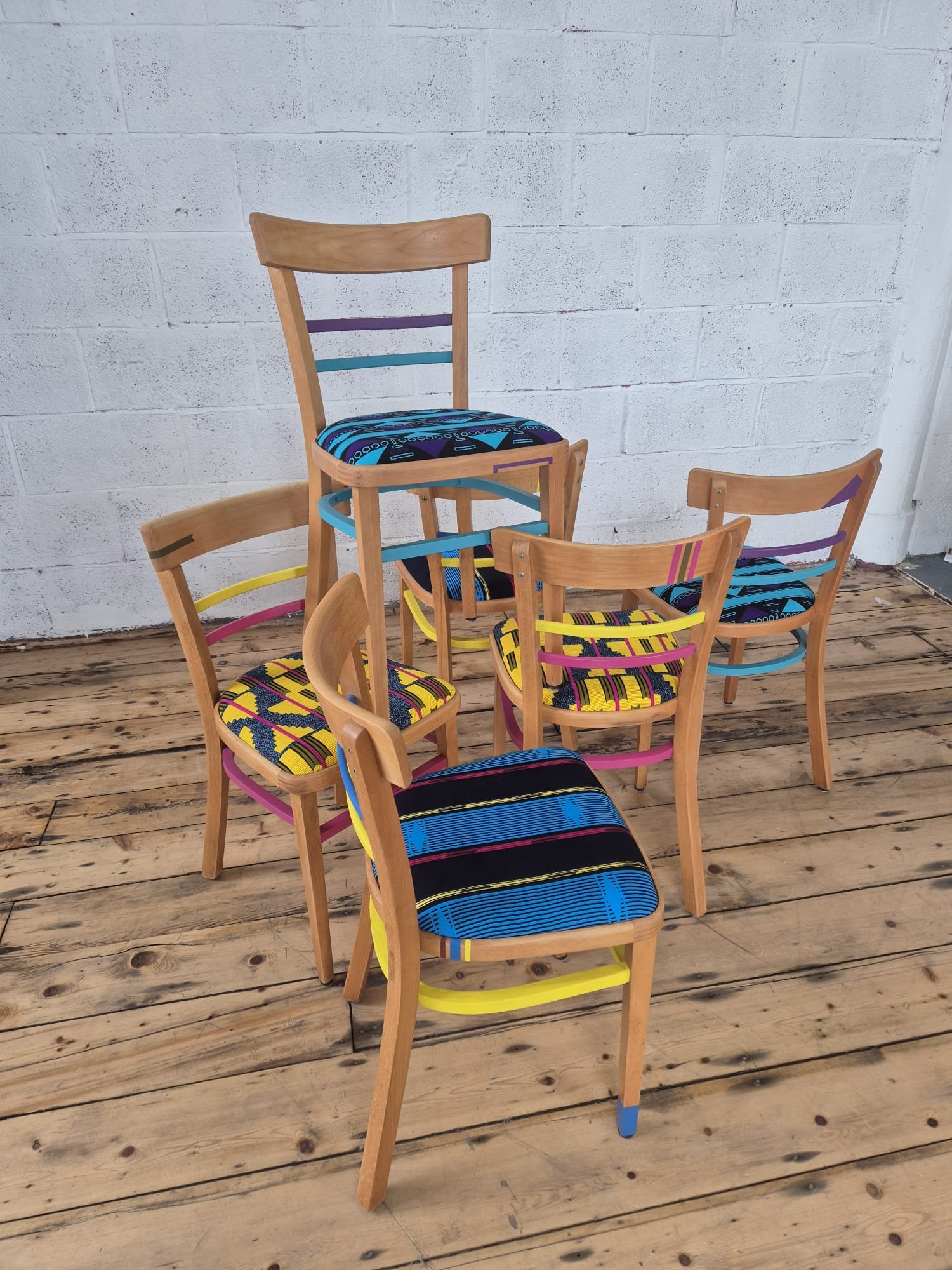 A group of five vintage wooden chairs with colorful patterned cushions arranged on a wooden floor against a white brick wall.
