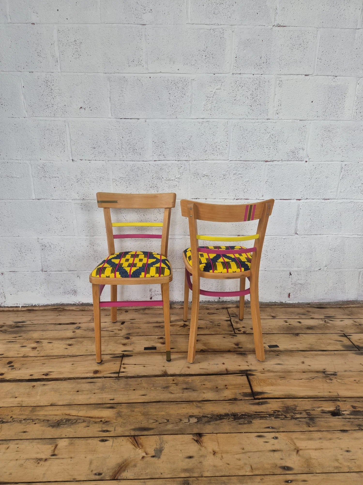 Two wooden chairs with colorful, patterned upholstery seats are positioned against a white brick wall on a wooden floor.