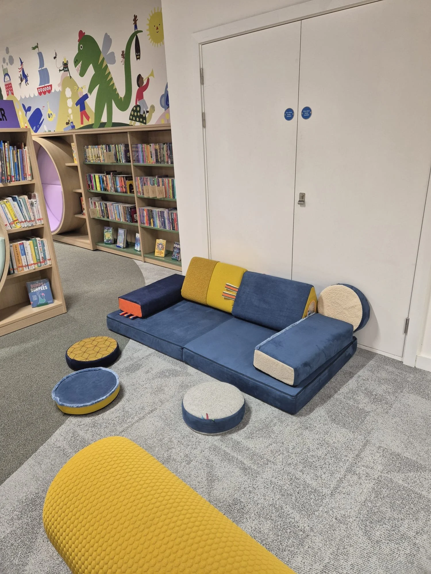 A cozy children's reading nook with a colorful, modern couch, round cushions, and a bookshelf filled with children's books. The wall has a vibrant mural of a dinosaur and whimsical characters. Based in Stockroom in Stockport Greater Manchester.