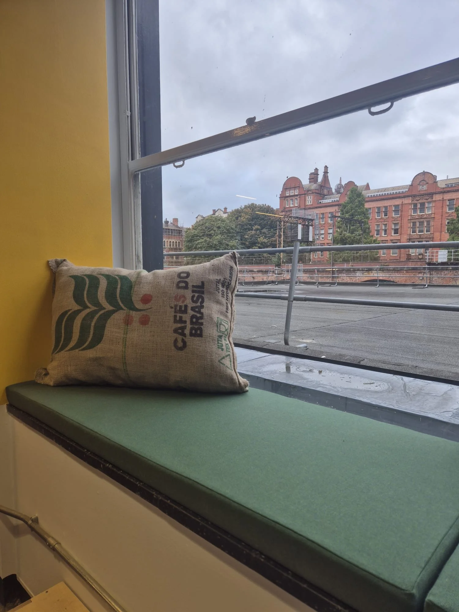 A window seat with a green cushion and a cushion made from a burlap coffee sack that says 'CAFÉ DO BRASIL' with a cityscape outside the window, including historic red-brick buildings and trees.