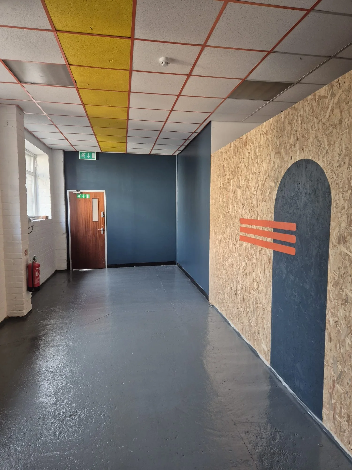 Interior hallway with a blue wall, white brick wall, and a wooden door. Ceiling with colorful tiles and a chalkboard wall with abstract art. Fire extinguisher on the white wall near windows.