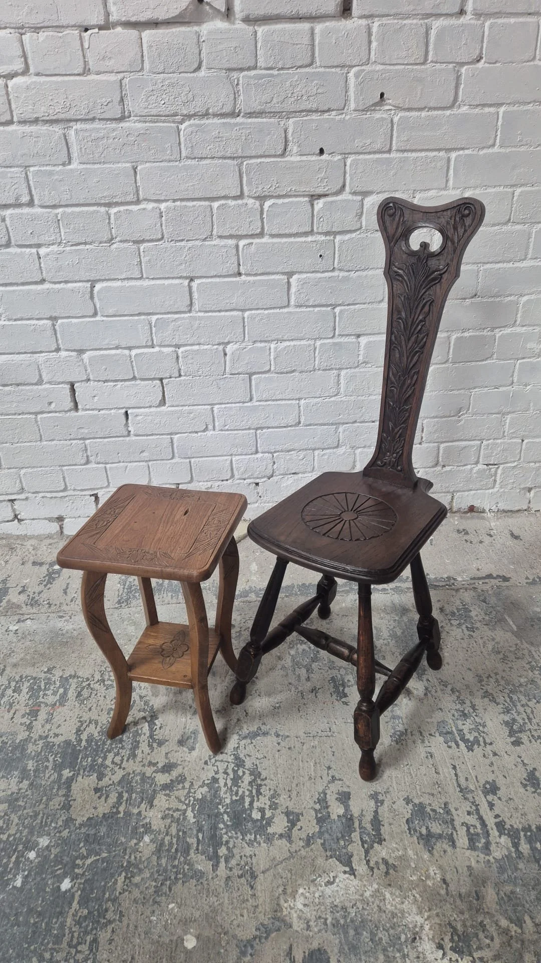 A vintage wooden high-back chair with carved detailing and a small wooden side table with a floral pattern on the top, both placed against a white brick wall on a distressed concrete floor.