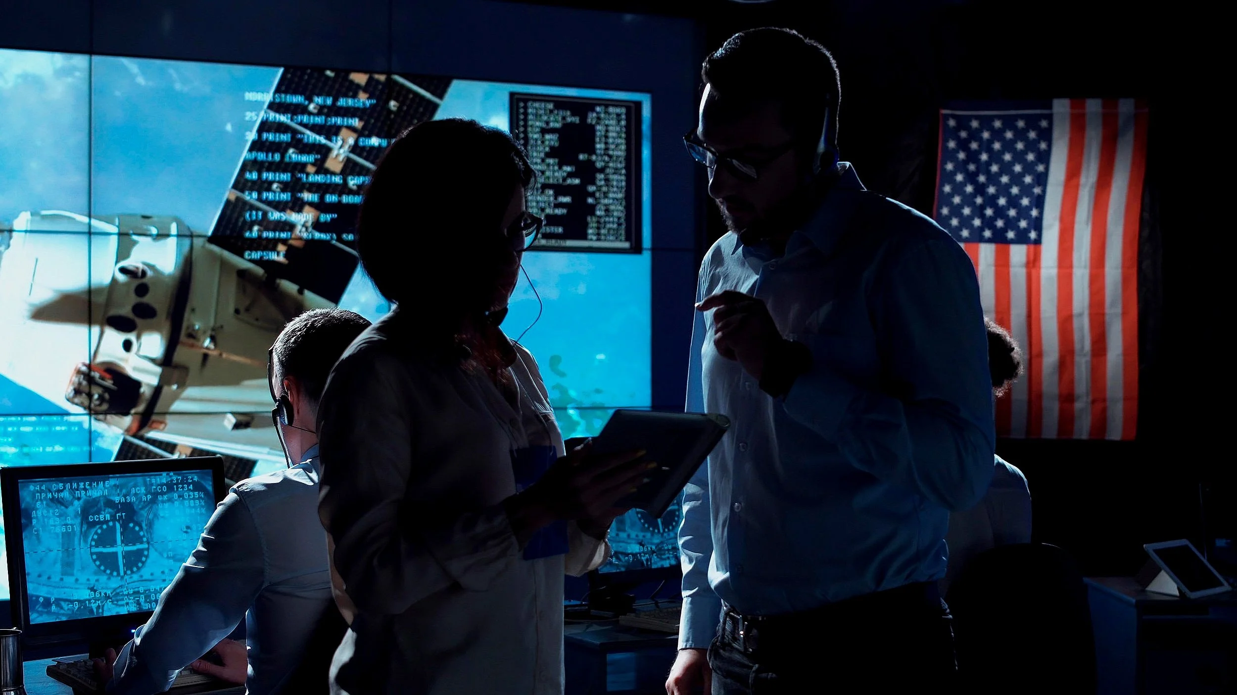 Silhouettes of four people working in a mission control room with large screens displaying space images and data, and an American flag on the wall.