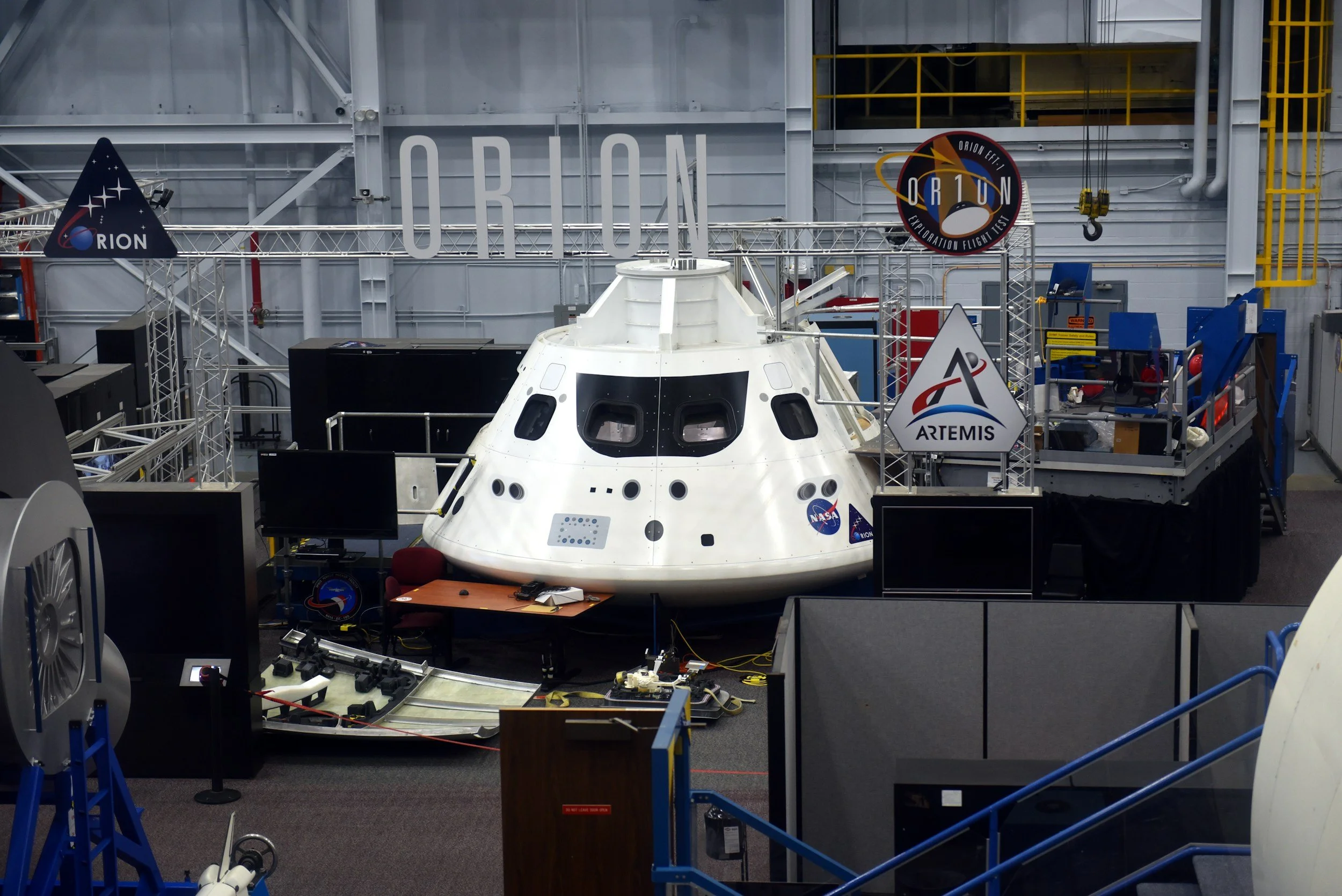 Interior of a space exploration facility with a spacecraft mock-up or capsule on display, surrounded by equipment, monitors, and NASA and Artemis logos.