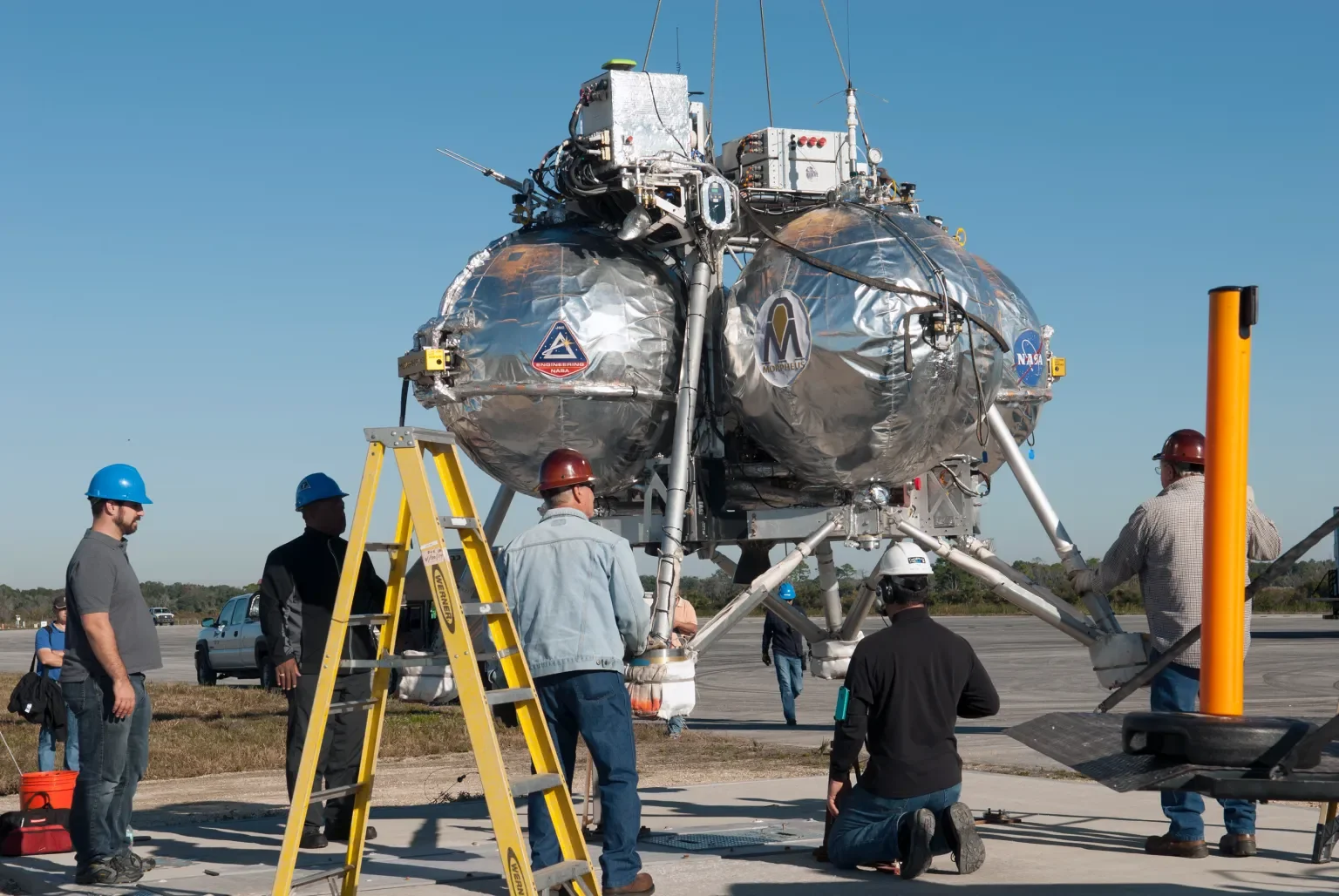 Spacecraft with technicians and engineers working on it on the ground, with a clear blue sky in the background.
