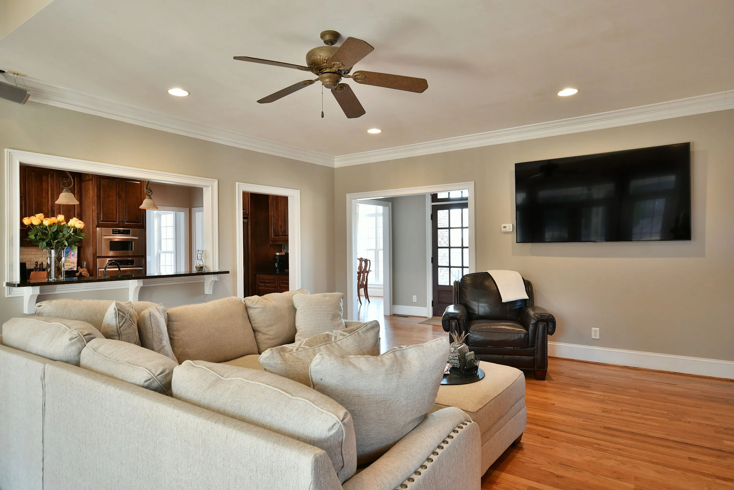 Living room with beige sofa, black armchair, wall-mounted TV, ceiling fan, hardwood floors, and open views into the kitchen and dining area. Home for sale in Greenville, SC. Master on Main Level. 4 Bedroom home for sale. Upscale Community. 