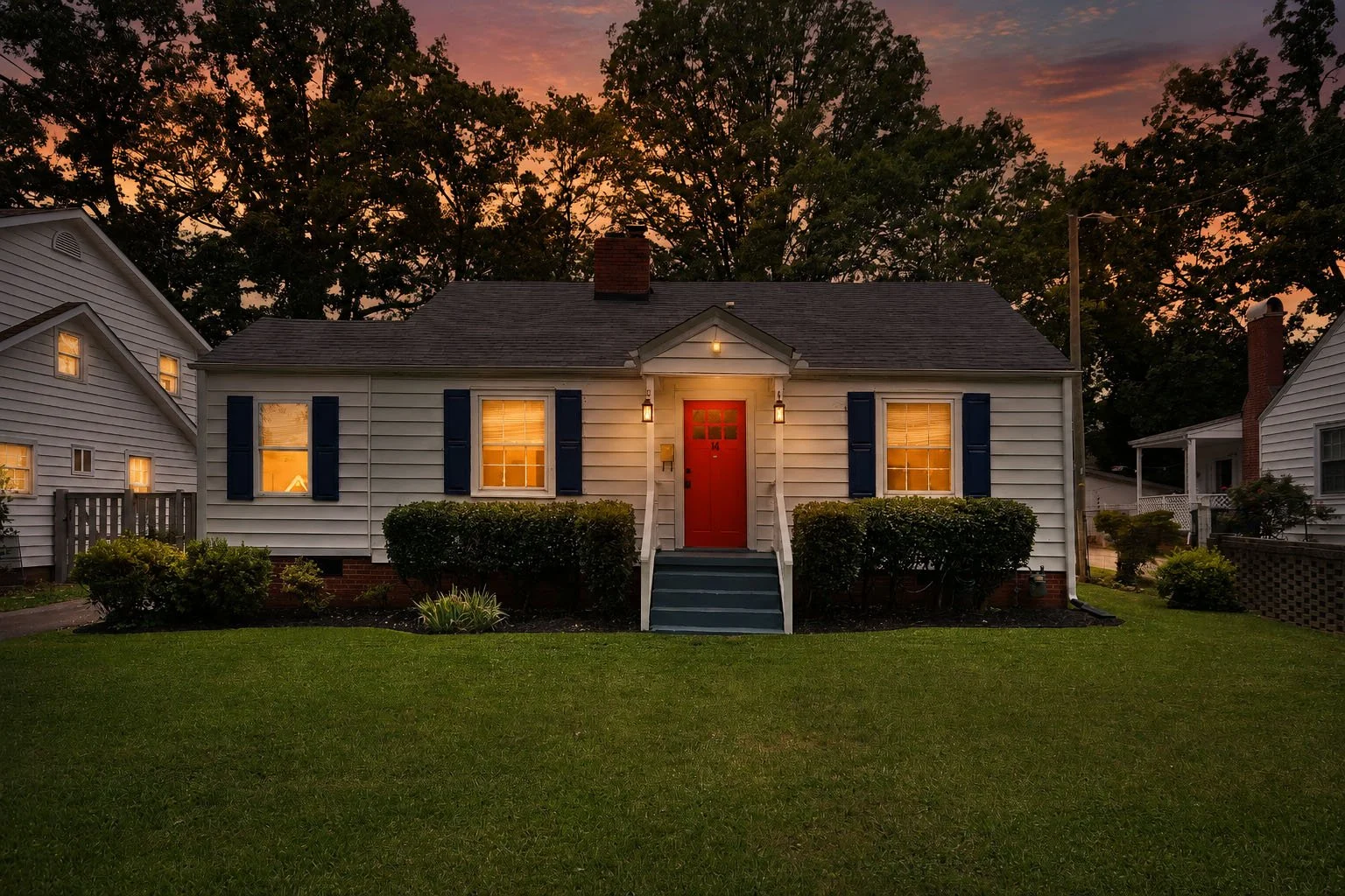 Front view of a white suburban house with black shutters and a red front door, illuminated by porch lights at sunset, with a well-maintained lawn and bushes in front.