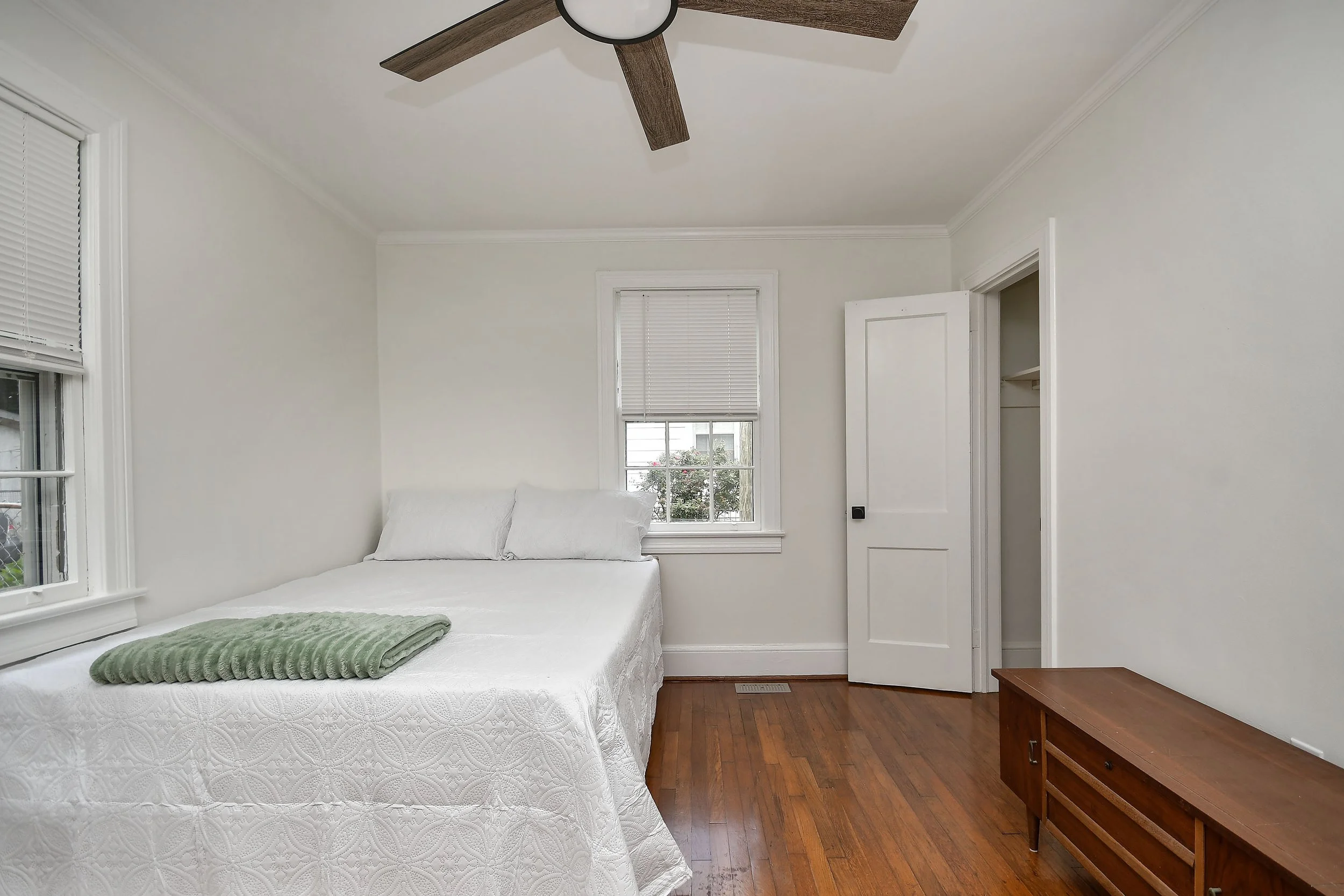 A bedroom with white walls, a bed with white bedding and pillows, a green blanket, a wooden bench at the foot of the bed, a window with blinds, a closed door, and a ceiling fan with wooden blades.