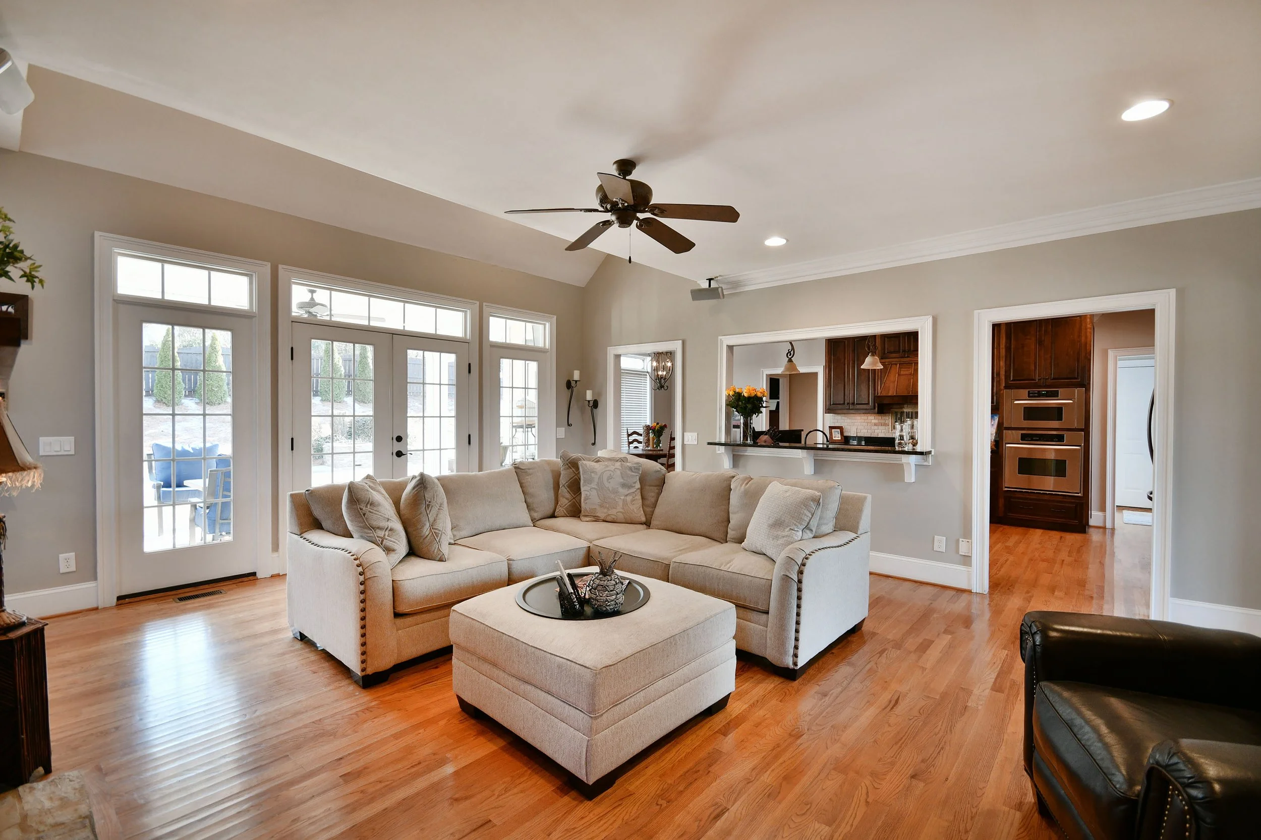 Living room with beige sectional sofa, ottoman with tray, black leather chair, hardwood floors, large windows and glass door to outside, ceiling fan, and view into kitchen with dark wood cabinets. Home for sale in Greenville, SC. Master on Main Level