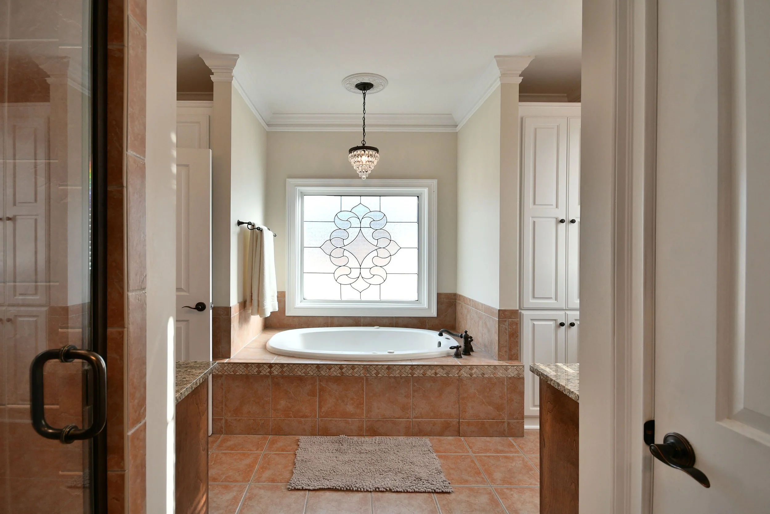 Bathroom with a bathtub under a window, decorated with a stained glass design, surrounded by white cabinetry and terracotta tile flooring. Home for sale in Greenville, SC. Master on Main Level. 4 Bedroom home for sale. Upscale Community. 