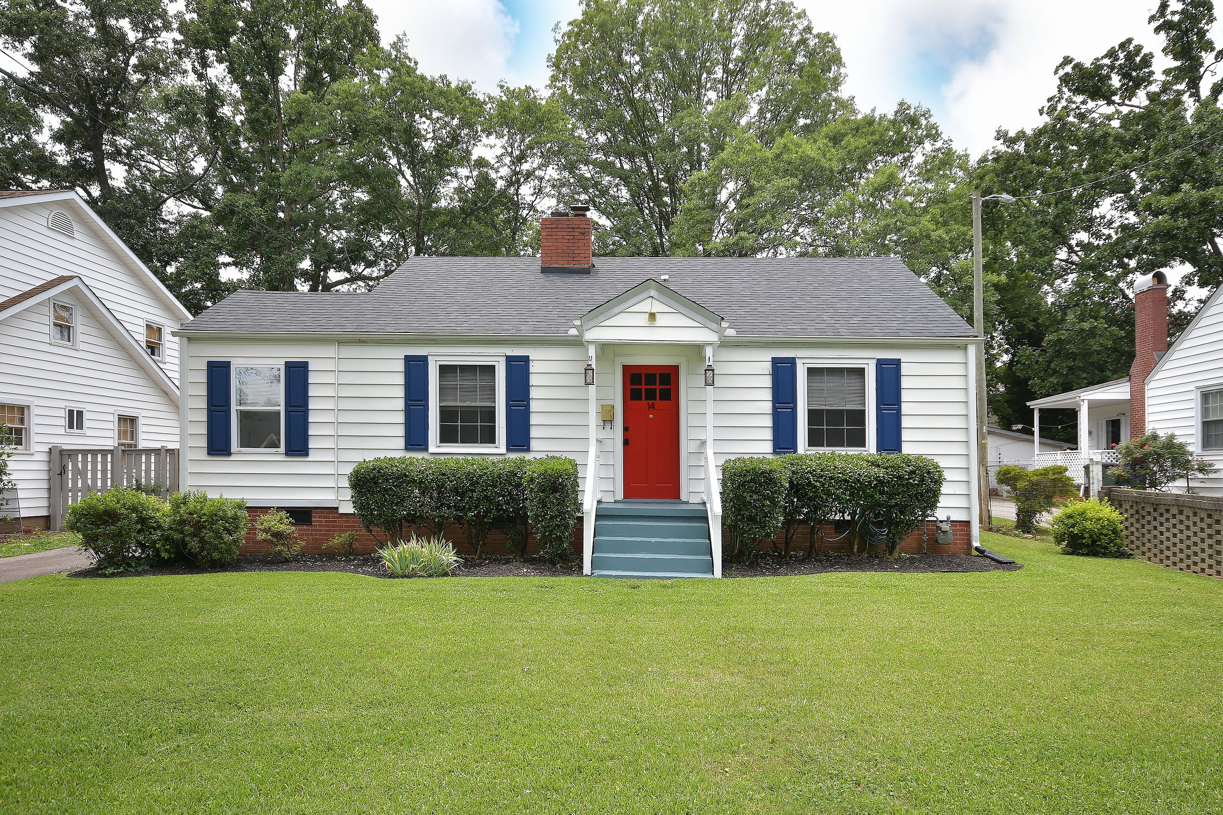 14 Tabor Street, a white house with blue shutters and a red door, surrounded by a well-maintained lawn and bushes, with large trees and neighboring houses in the background.