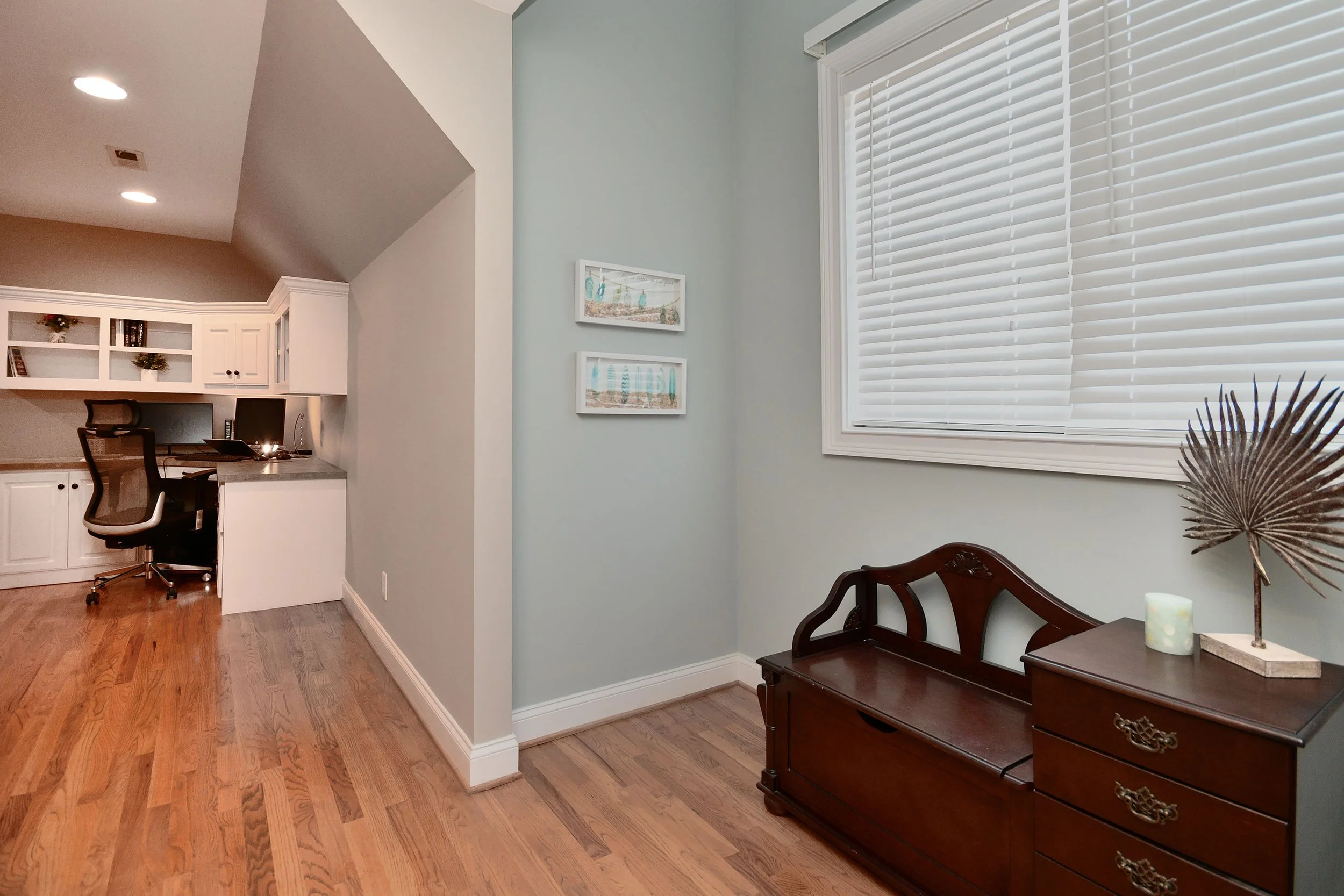 A cozy corner of a room with a wooden bench, a chest of drawers with decorative items, and a window with blinds, adjacent to a workspace with a desk and chair, and wall art. Home for sale in Greenville, SC. Master on Main Level. 4 Bedroom home 