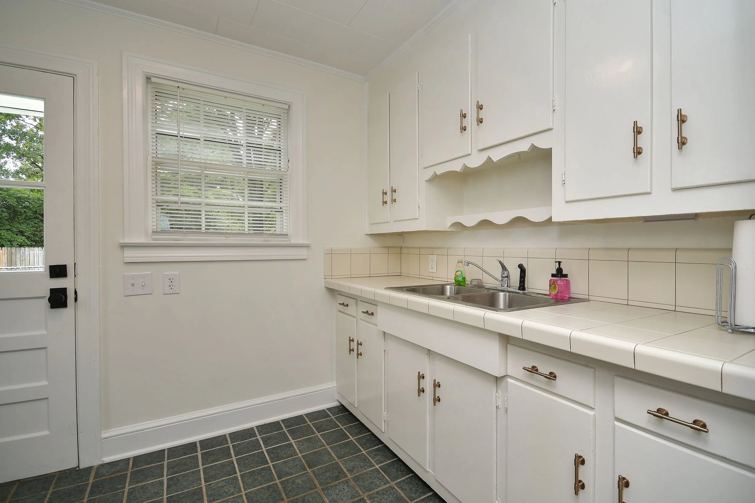A white kitchen with a window covered by white blinds, a white tiled countertop with a sink, drying rack, soap dispenser, and a paper towel roll. White cabinets with brass handles are above and below the countertop. The floor has dark square tiles.