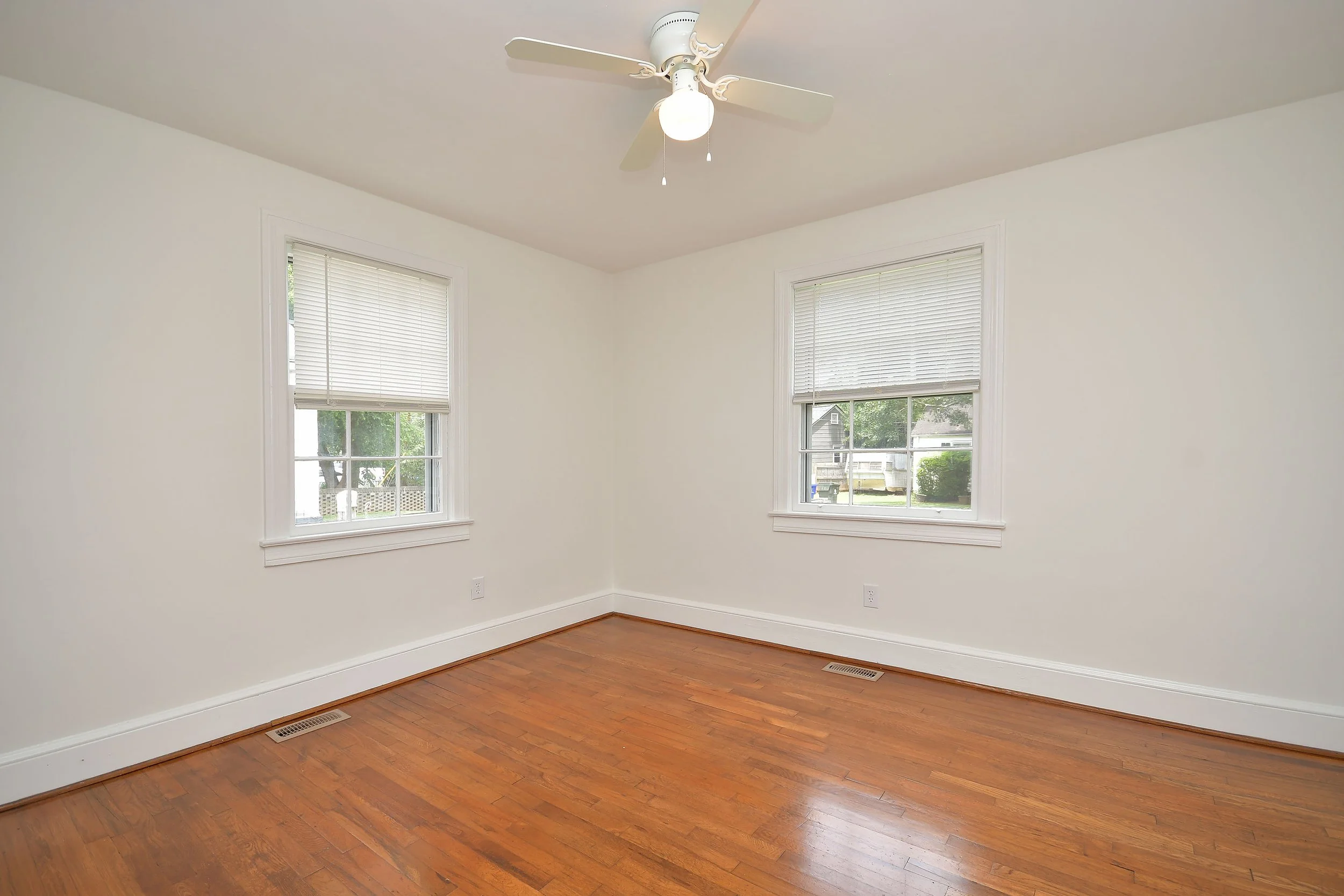Empty room with hardwood floor, white walls, two windows with blinds, ceiling fan with light, and vents on the floor.