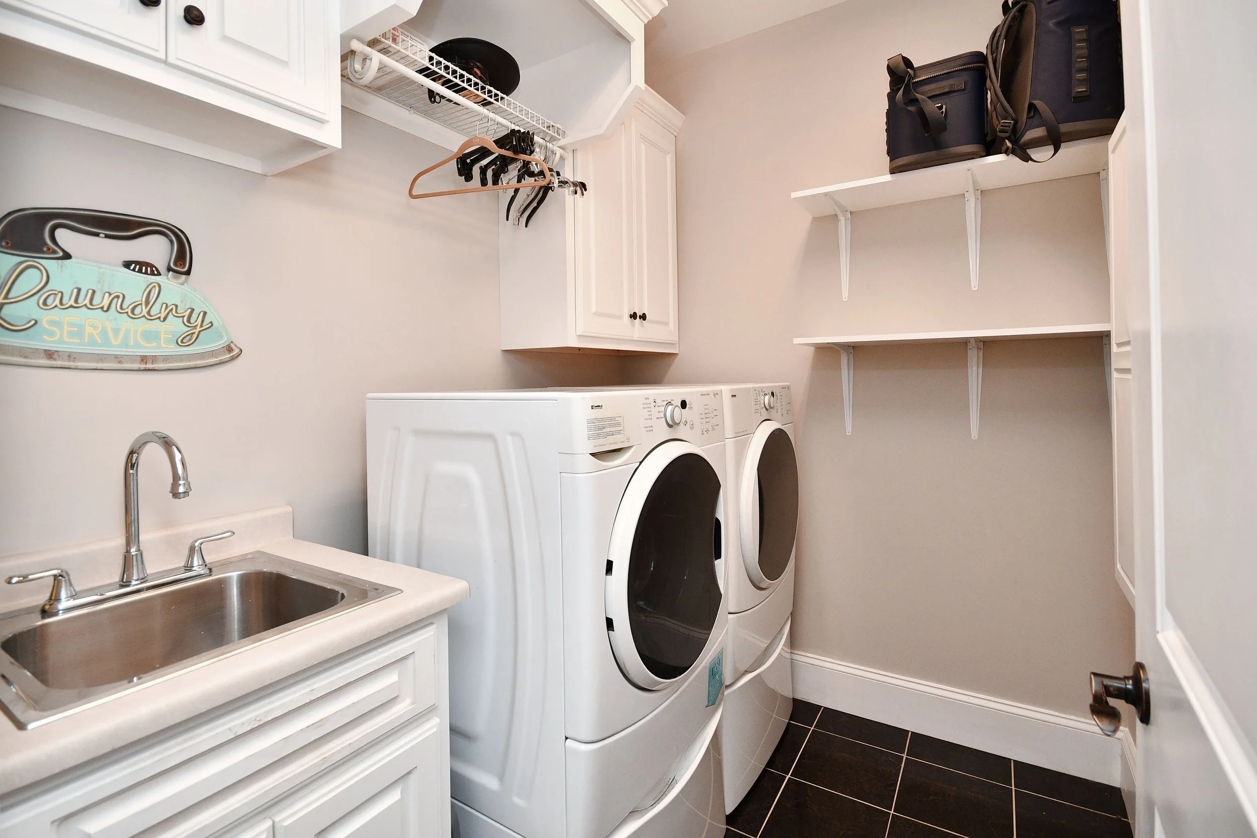 Laundry room with white washer and dryer, a small sink with a chrome faucet, white cabinets, black and white tiled flooring, and shelves holding bags and a plaid box. Home for sale in Greenville, SC. Master on Main Level. 4 Bedroom home for sale. 