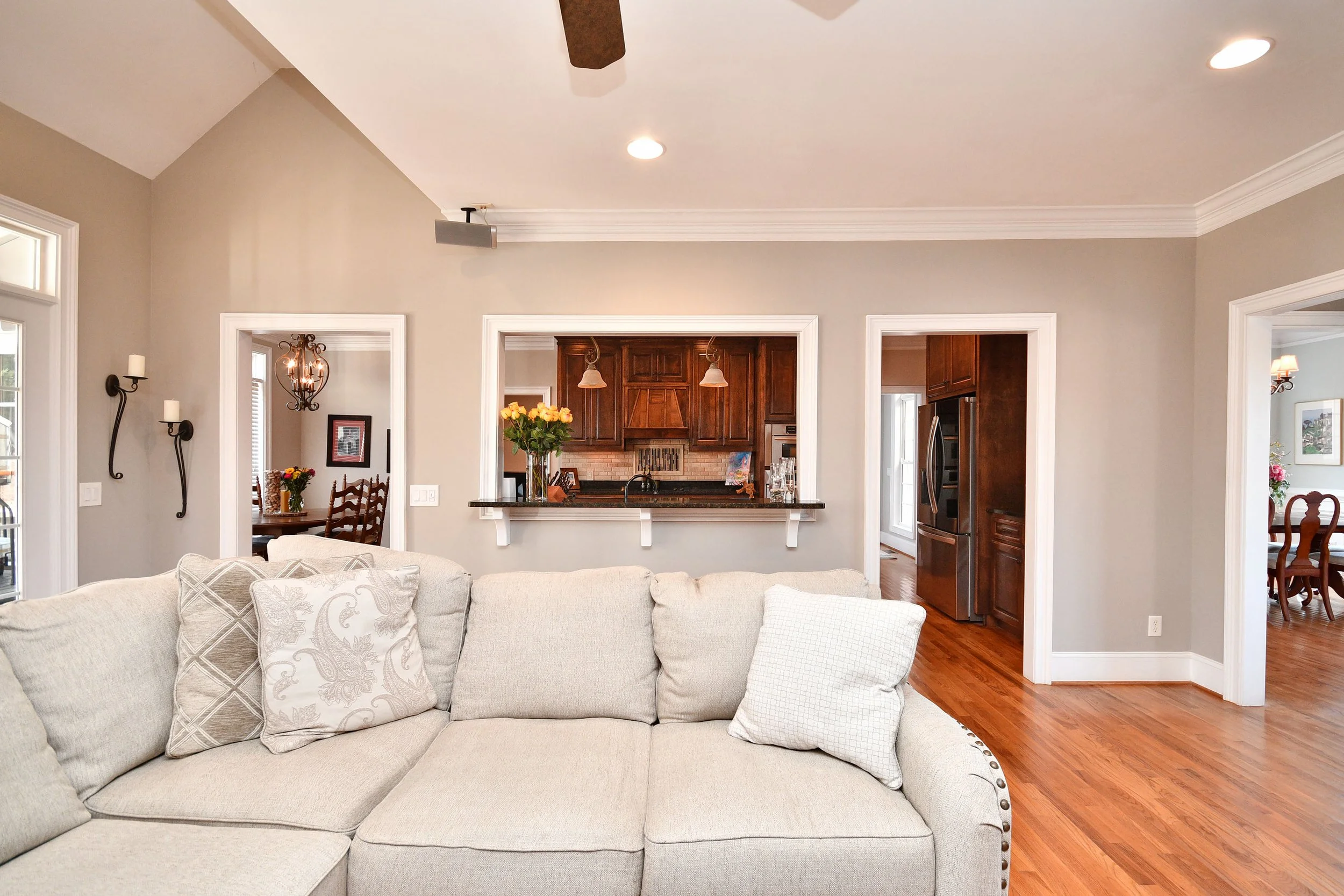 Living room with beige sofa, decorative pillows, hardwood floors, open kitchen with dark wood cabinets, flowers, and dining area in the background. Home for sale in Greenville, SC. Master on Main Level. 4 Bedroom home for sale. Upscale Community. 