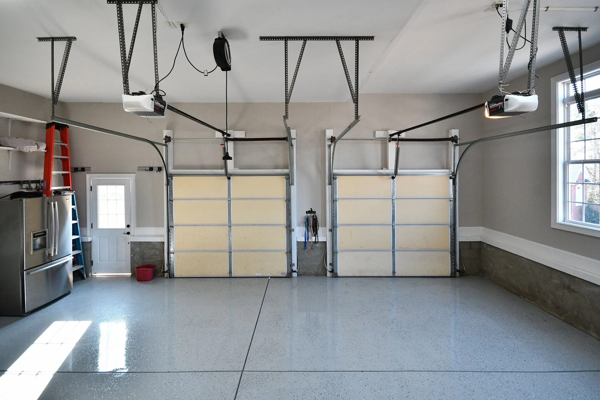 Empty two-car garage with beige rolling doors, a stainless steel refrigerator, a ladder, and garage door openers mounted on the ceiling. Home for sale in Greenville, SC. Master on Main Level. 4 Bedroom home for sale. Upscale Community. 