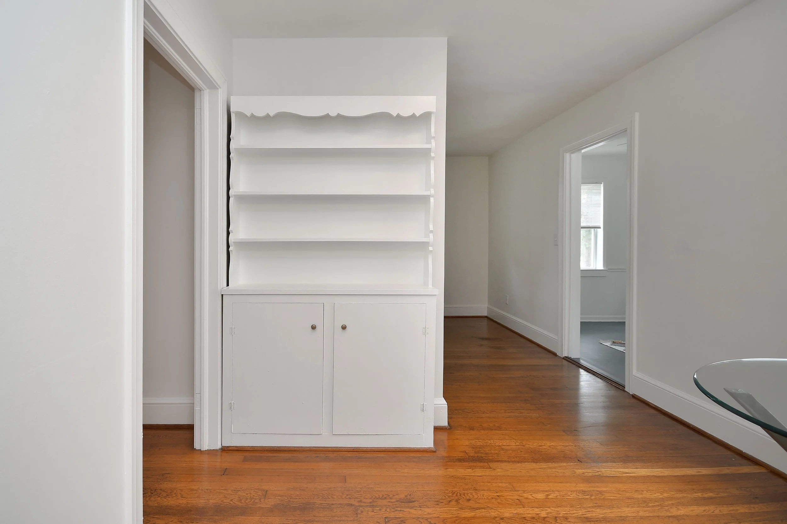 Interior view of a room with wooden floors, white walls, and an open doorway. A white built-in cabinet with shelves is against the wall.
