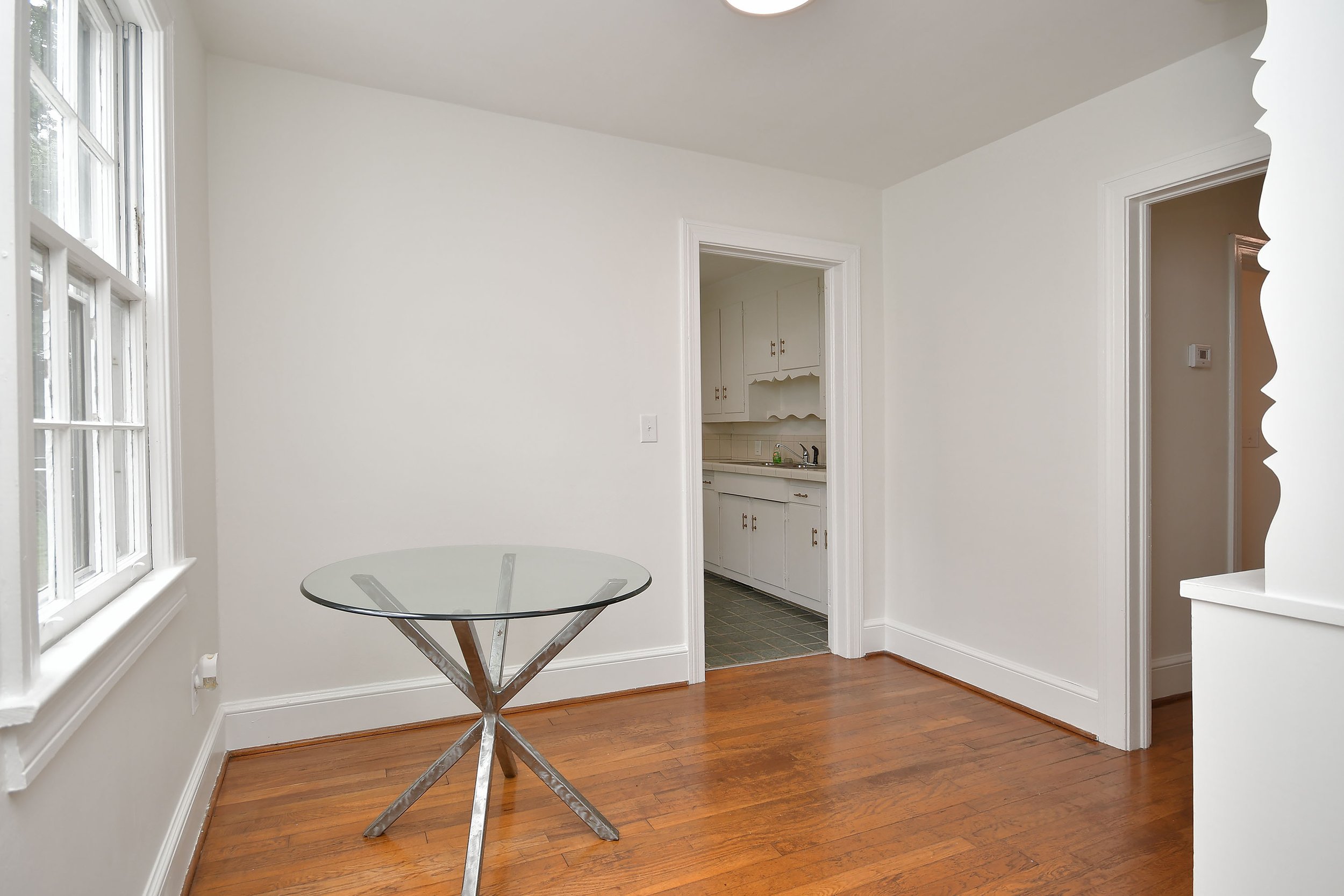 A dining area with a glass table, wooden floor, large window, and views into a kitchen with white cabinets.