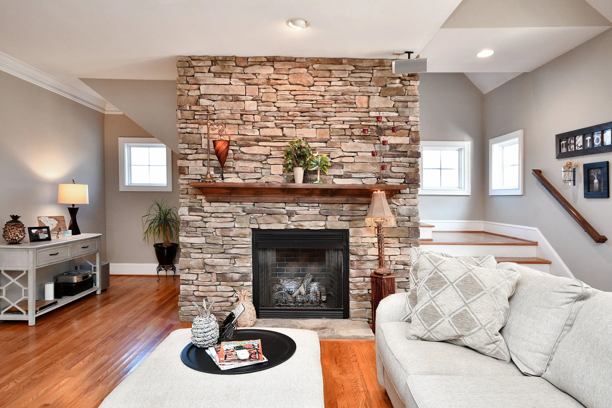 Living room with a stone fireplace, wooden mantel, beige sofa, and hardwood flooring, decorated with plants, lamps, and wall art. Home for sale in Greenville, SC. Master on Main Level. 4 Bedroom home for sale. Upscale Community. 
