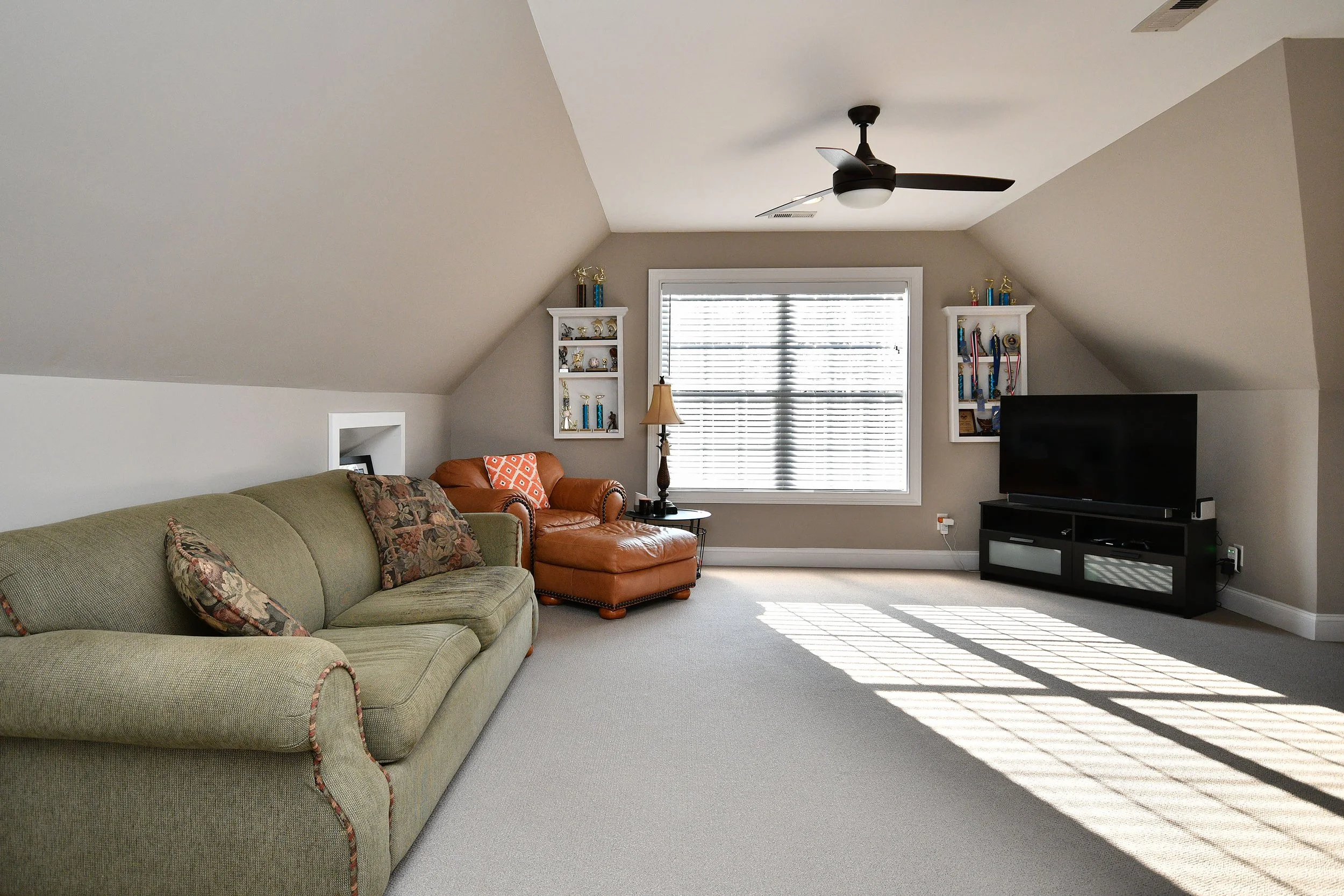 Living room with sloped ceilings, beige walls, a large window with blinds, a ceiling fan, a green sofa with patterned pillows, a brown leather armchair with an orange pillow, a small side table with a lamp, and a black TV stand with a flat screen TV.