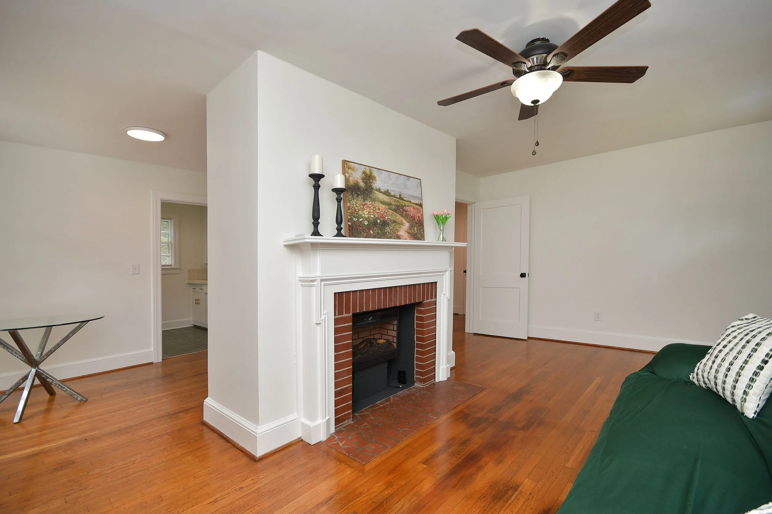 Living room with a white fireplace, wooden floor, ceiling fan with lights, painting of a landscape, pink tulips in a glass vase, and green furniture.