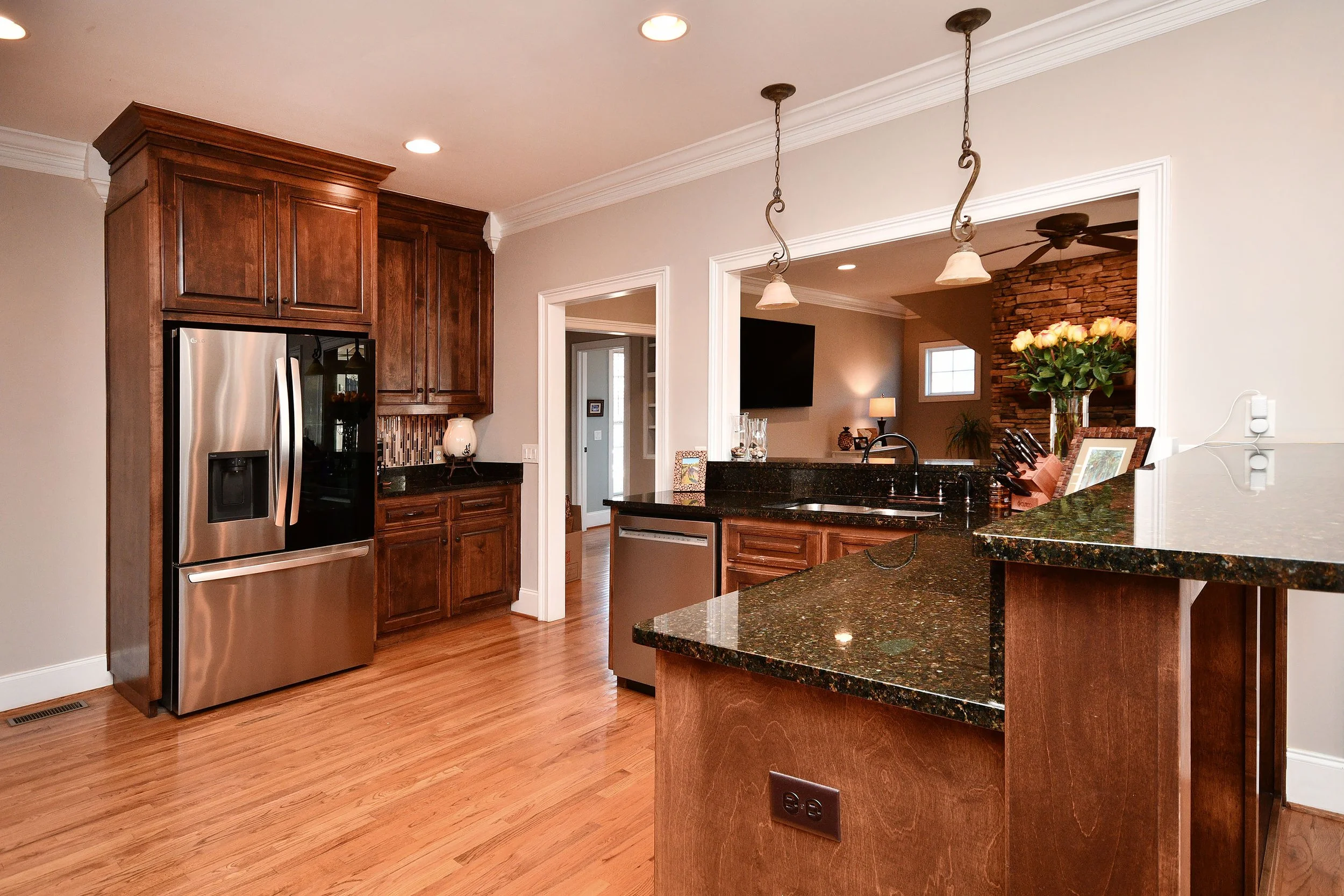 A modern kitchen with wooden cabinets, stainless steel refrigerator, black granite countertops, and hardwood floors, with a view into the living room. Home for sale in Greenville, SC. Master on Main Level. 4 Bedroom home for sale. Upscale Community. 