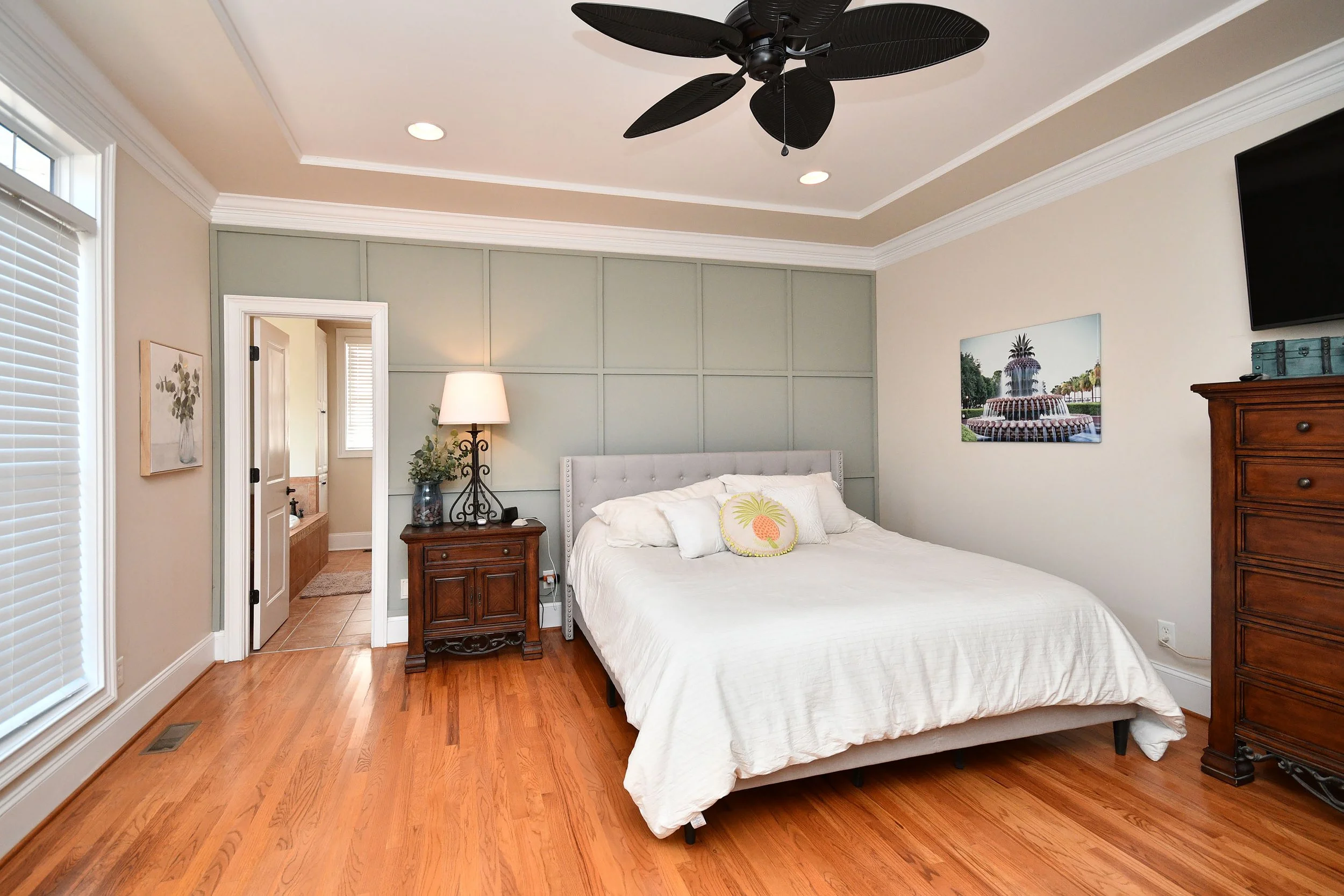 A bedroom with a white bed, wooden furniture, a ceiling fan, and wall art, with an en suite bathroom visible through an open door. Home for sale in Greenville, SC. Master on Main Level. 4 Bedroom home for sale. Upscale Community. 