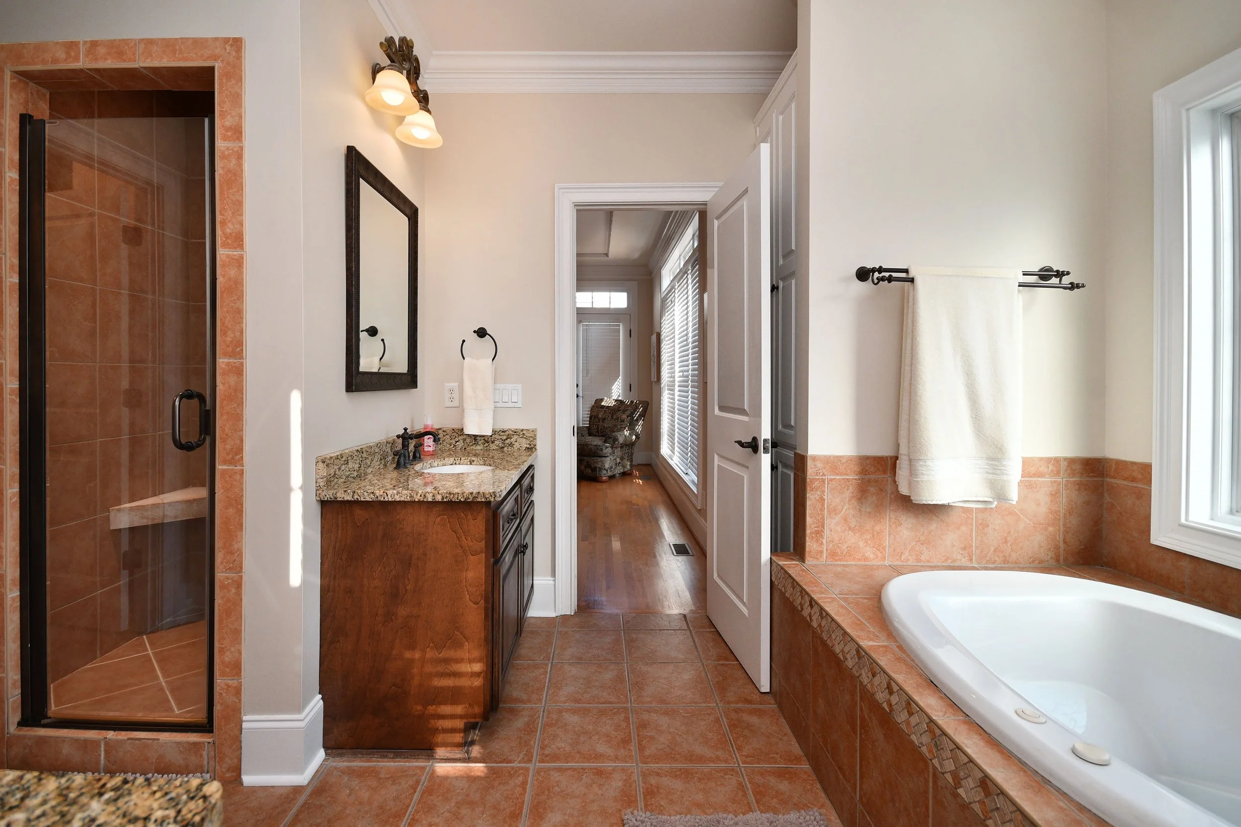 Bathroom with walk-in shower, vanity with granite countertop, and bathtub, featuring orange tiles, white walls, and a view into an adjacent room with wooden floor and large windows. Home for sale in Greenville, SC. Master on Main Level. 4 Bedrooms