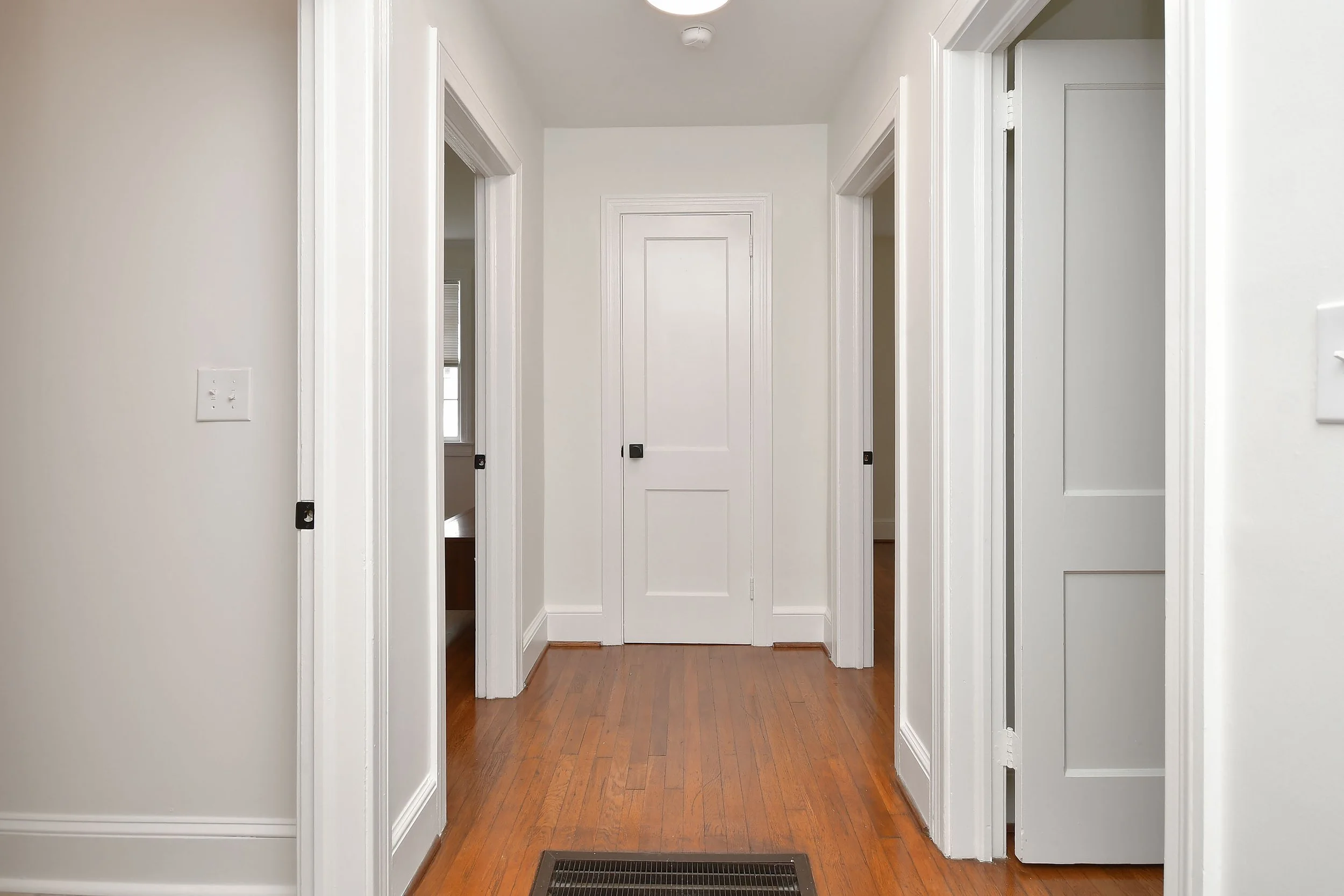 View of a hallway with white walls, wooden flooring, multiple doorways, and a vent cover on the floor.