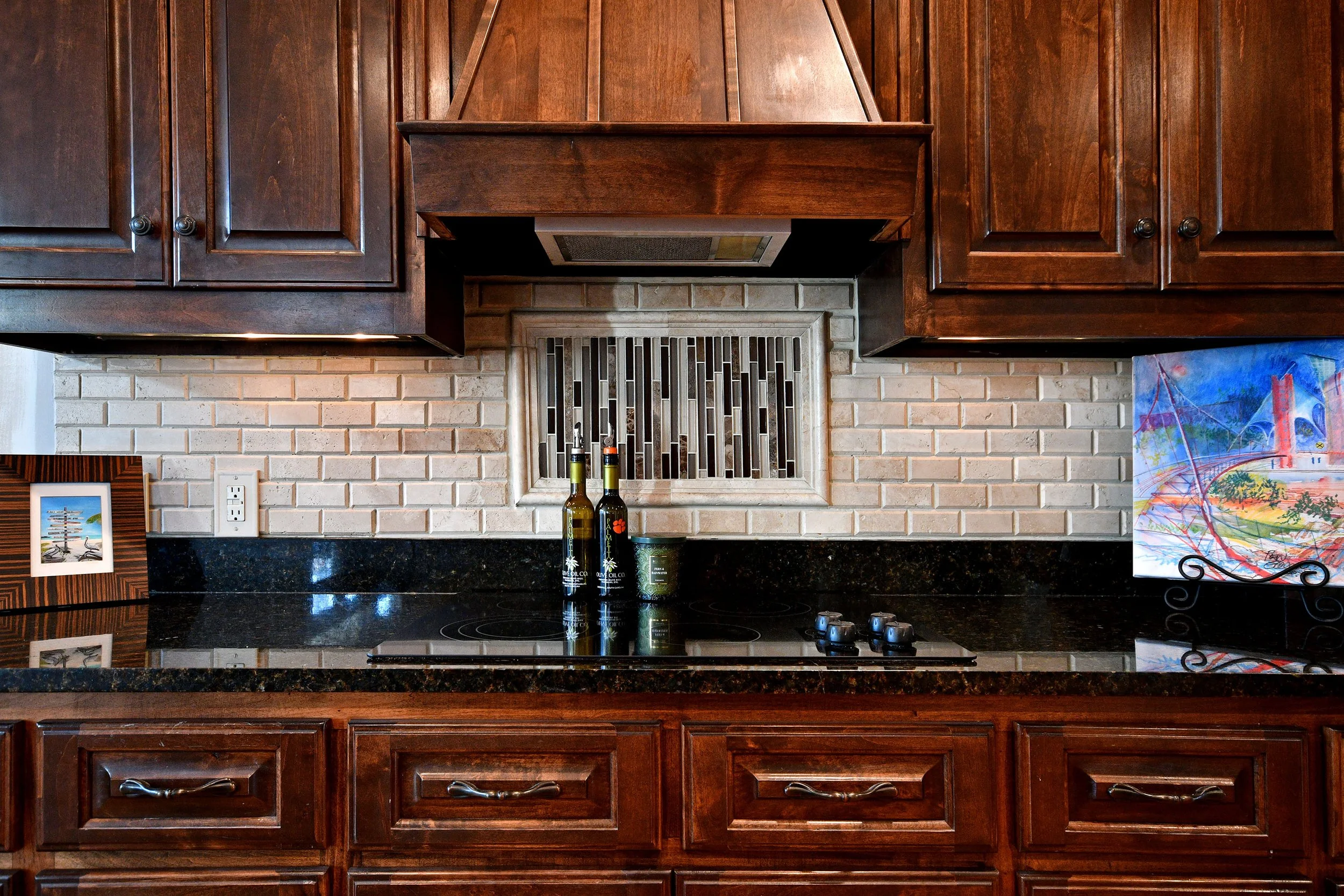 Wooden kitchen cabinets with black countertops and brick backsplash, featuring a small framed picture and colorful painting on display, with bottles and a container on the counter. Home for sale in Greenville, SC. Master on Main Level. 4 Bedroom home