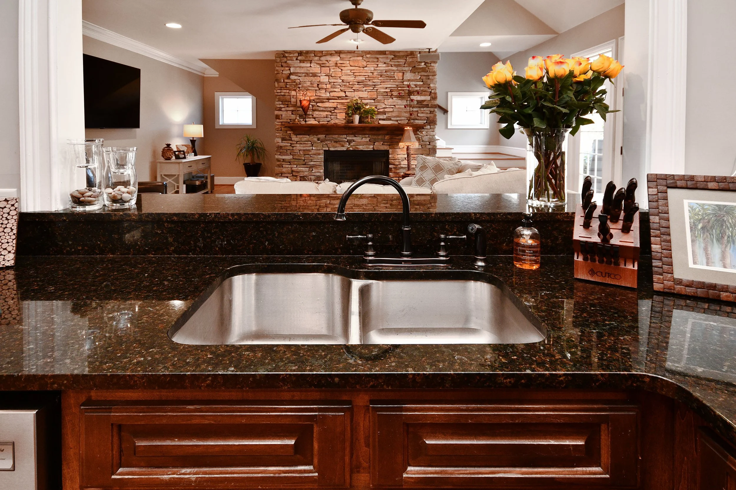 Kitchen view with a granite countertop, a stainless steel sink, a faucet, and a vase of yellow roses, with a living room area with a stone fireplace in the background. Home for sale in Greenville, SC. Master on Main Level. 4 Bedroom home for sale. 