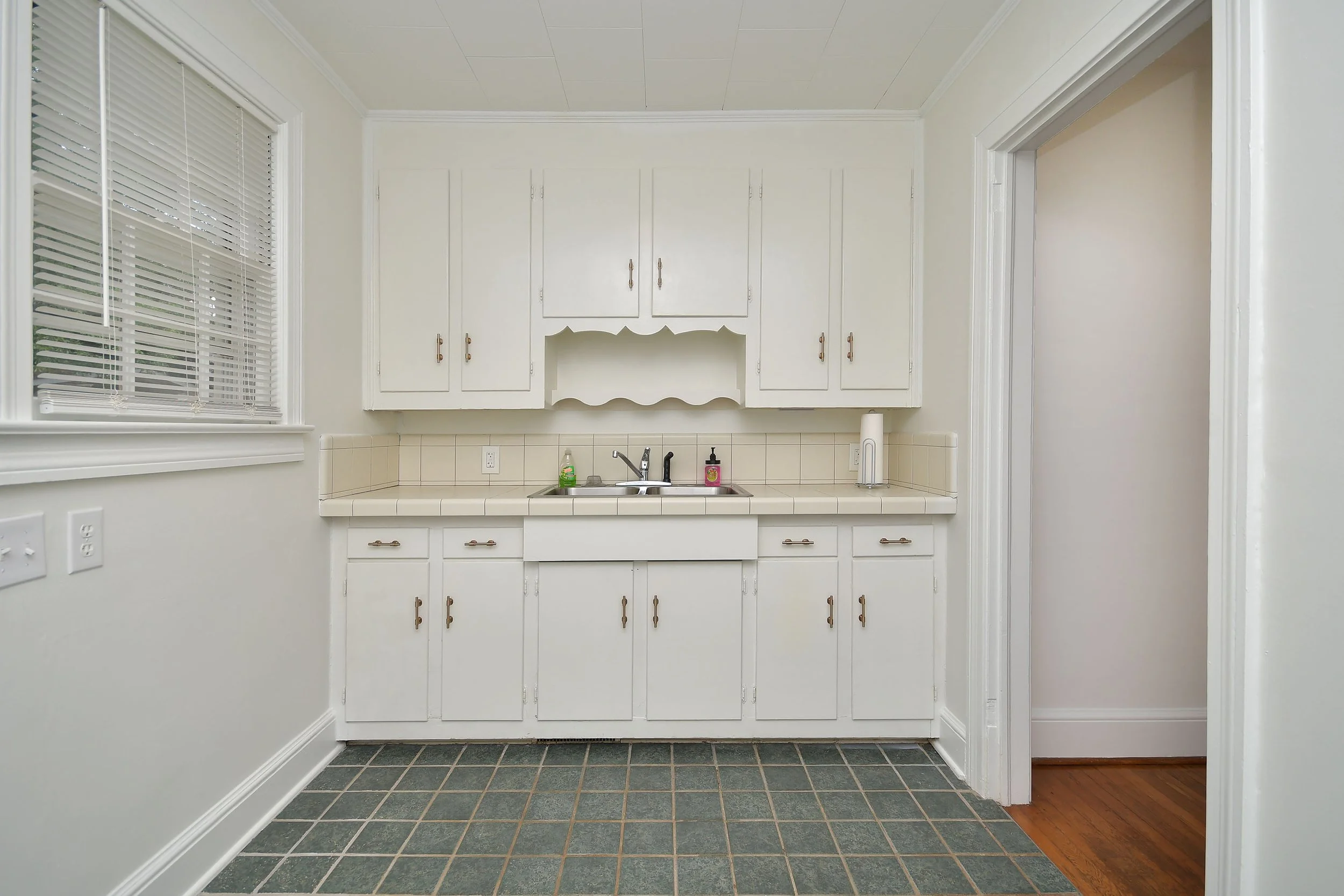 A kitchen with white cabinets, a stainless steel sink, window with white blinds, tiled countertop, and a tiled floor, with an adjacent room with wooden flooring.