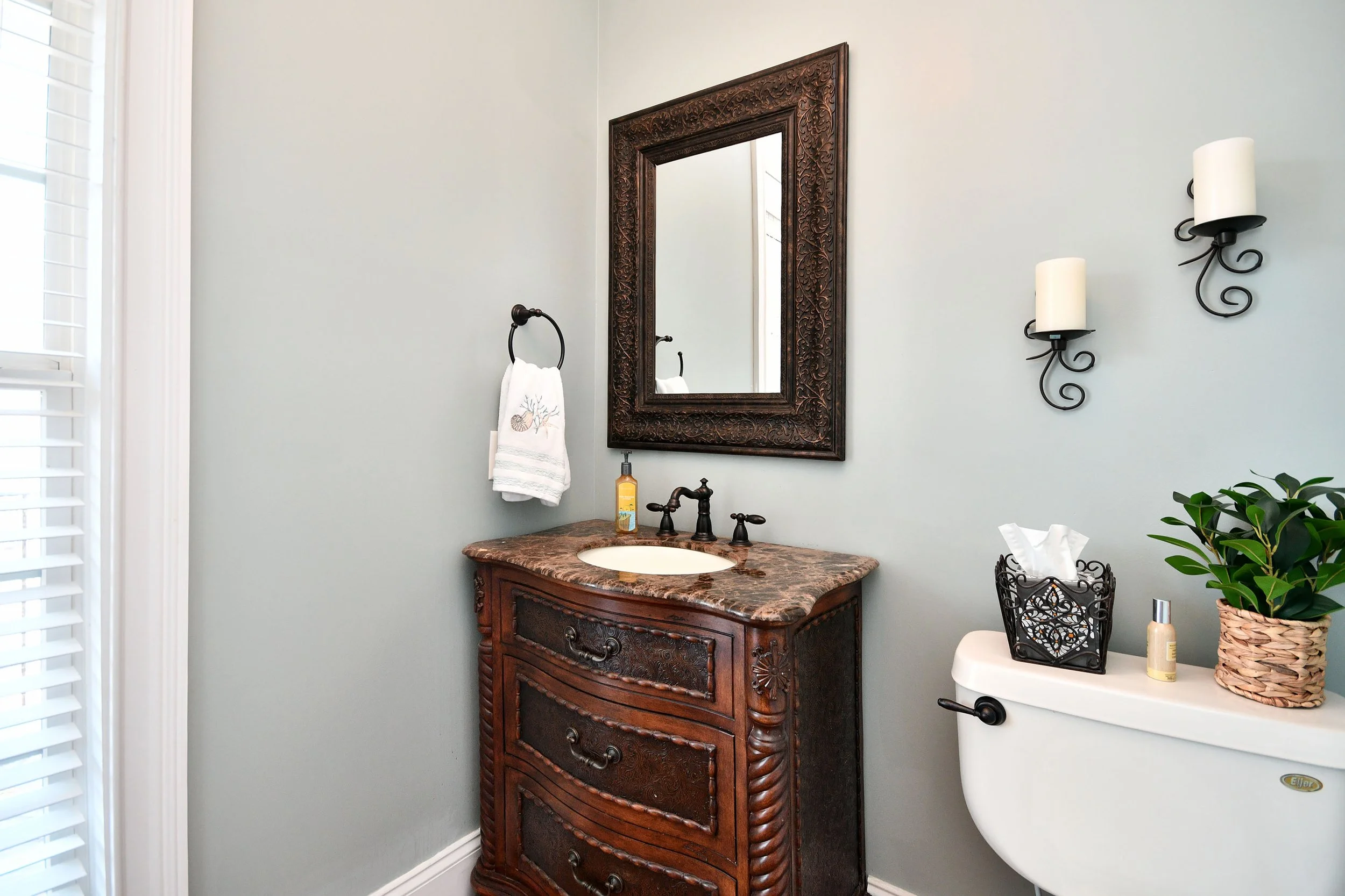 Bathroom with antique wooden vanity, marble countertop, and decorative mirror; wall-mounted candle sconces; toilet with tissue box and plant on top. Home for sale in Greenville, SC. Master on Main Level. 4 Bedroom home for sale. Upscale Community. 