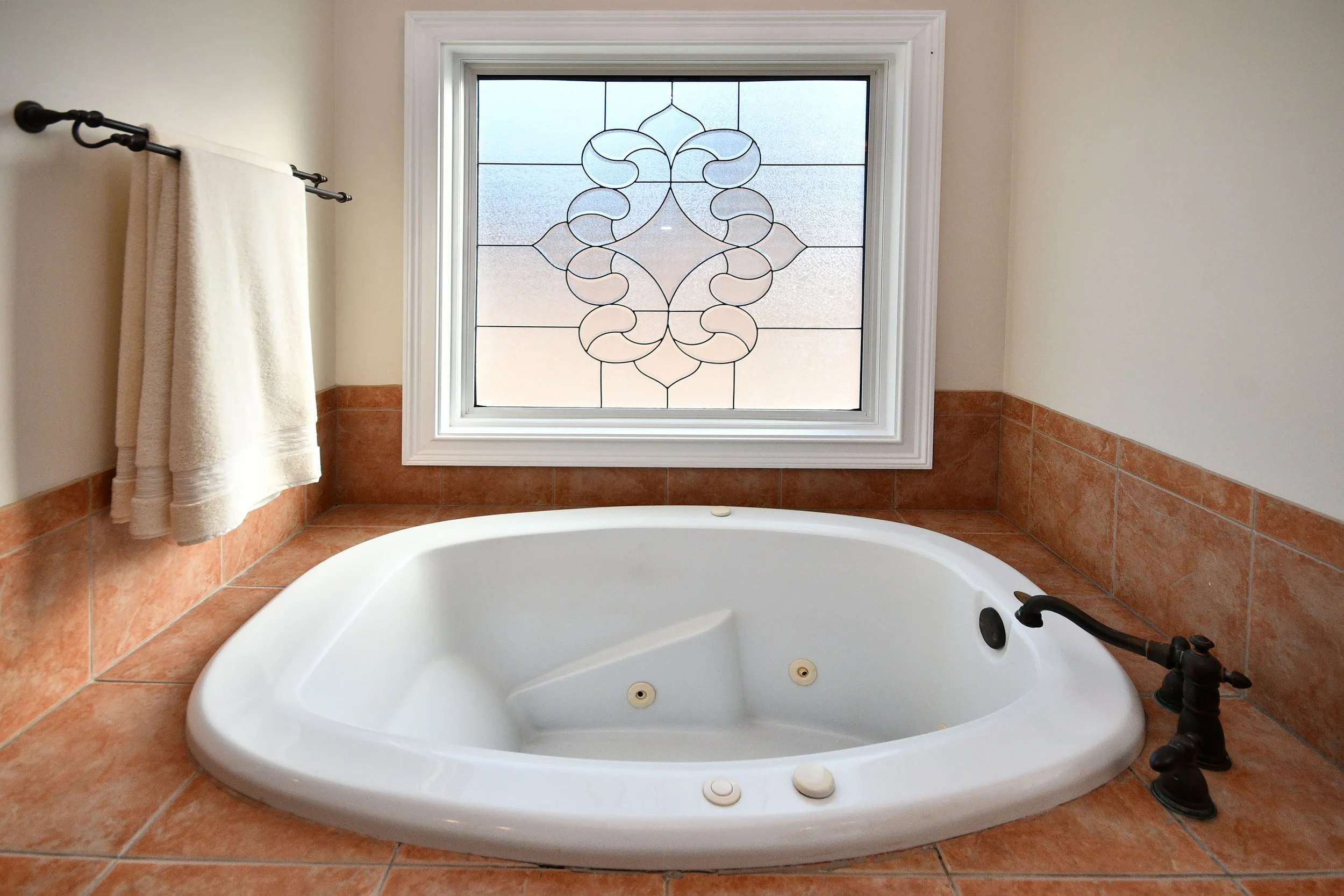 A bathtub with a white interior, brownish-red tile surrounding it, a black faucet on the right side, and a window with decorative stained glass design above. Home for sale in Greenville, SC. Master on Main Level. 4 Bedroom home for sale. Upscale.