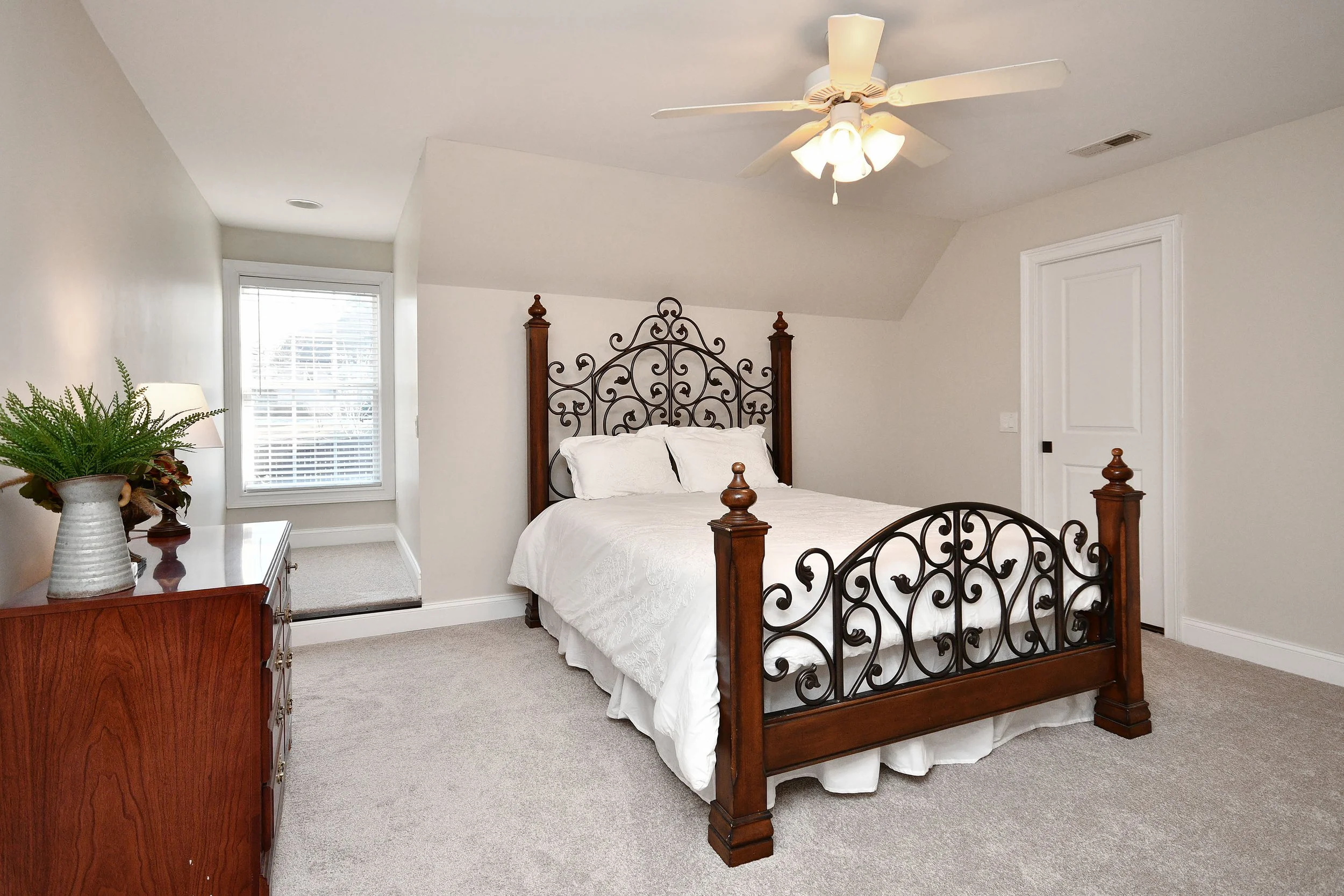 A bedroom with a four-poster bed with ornate metal headboard and footboard, white bedding, a wooden dresser with a plant, a window with blinds, a ceiling fan, and a carpeted floor. Home for sale in Greenville, SC. Master on Main Level. 4 Bedroom home