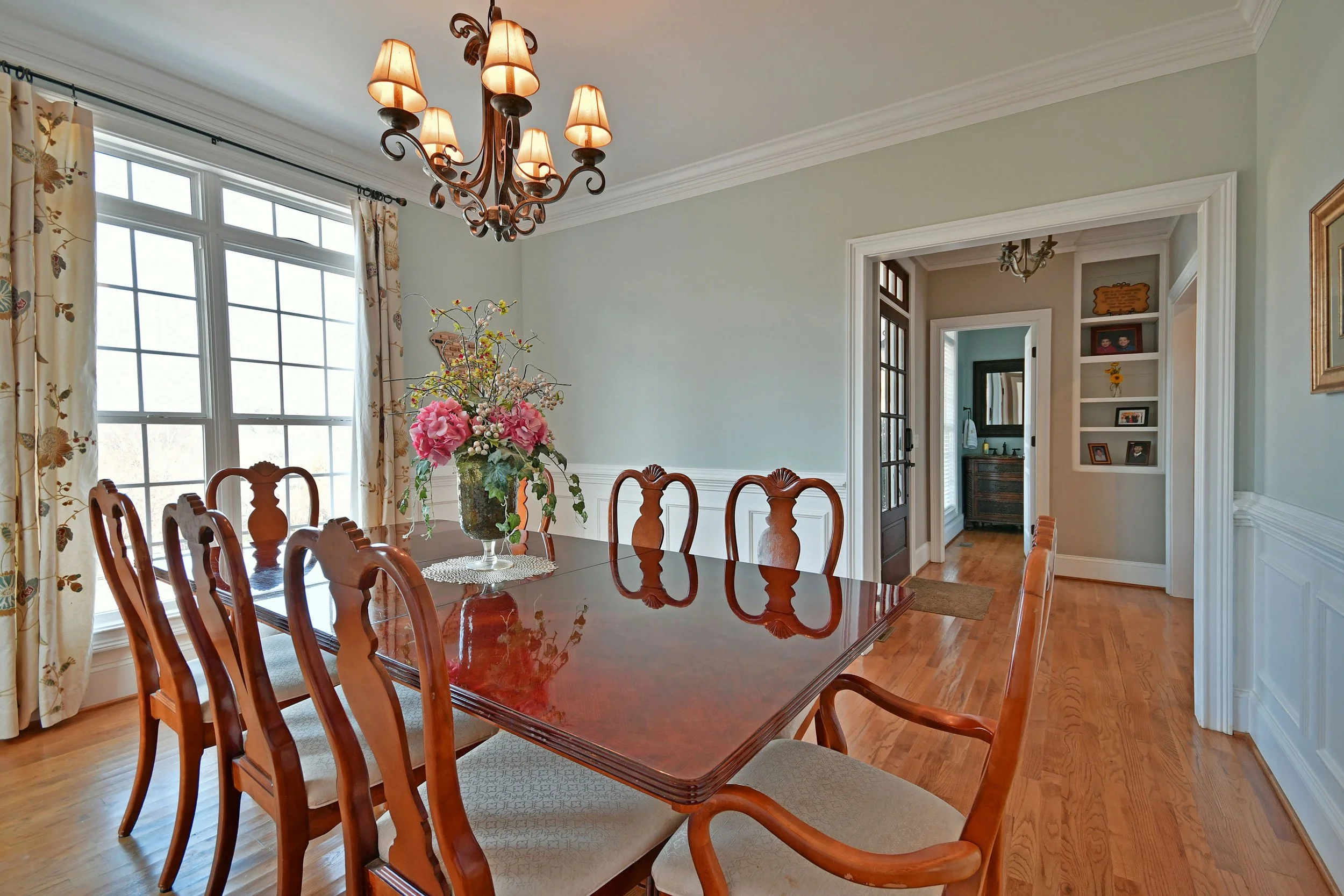 A dining room with a wooden table, matching chairs, a large floral centerpiece, a chandelier, and hardwood floors. There is a doorway leading to another room and built-in shelves on the wall. Home for sale in Greenville, SC. Master on Main.