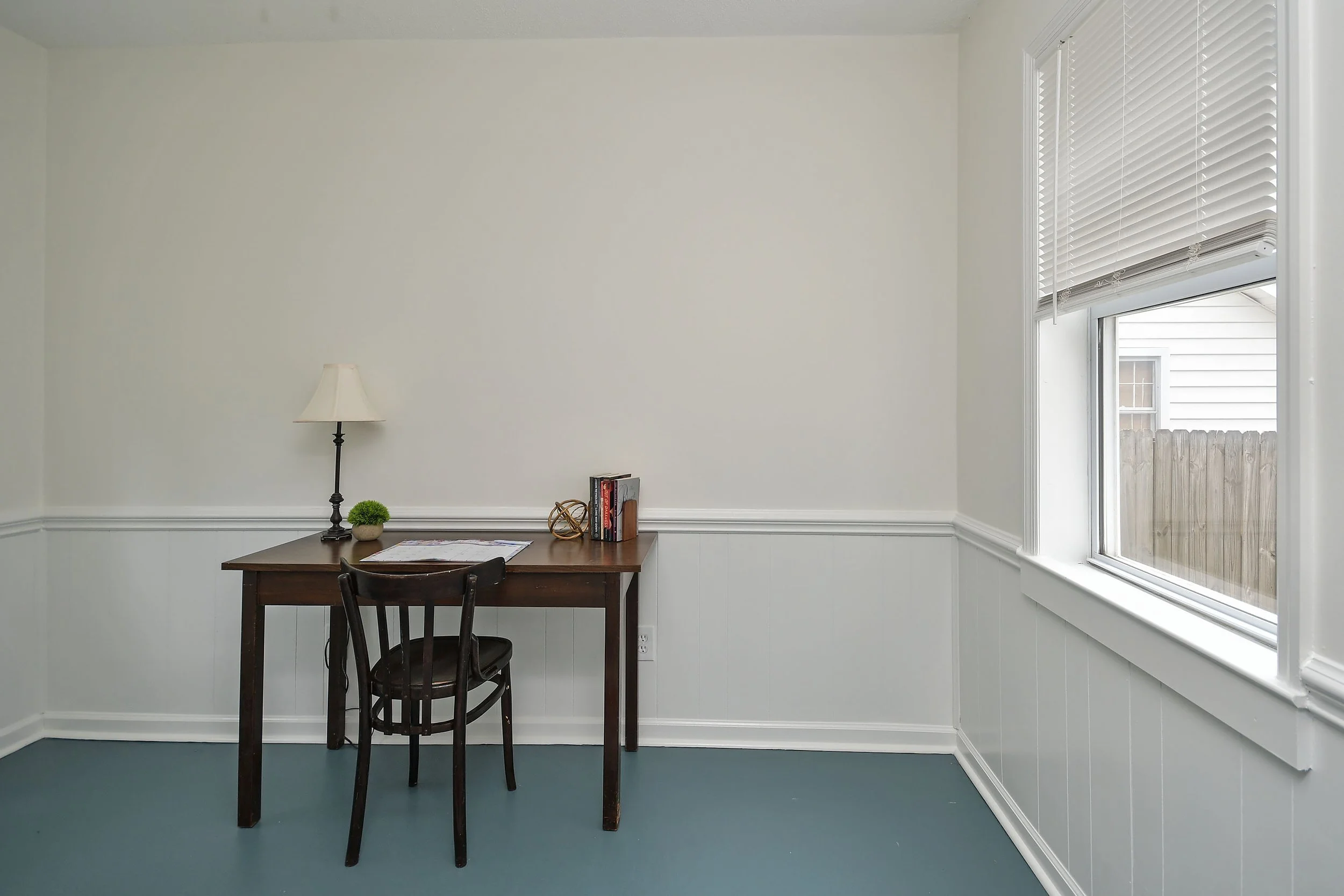 A small room with white walls and a large window with white blinds. Against the wall, there is a small wooden table with a black chair, a lamp, a small potted plant, and some books and papers.