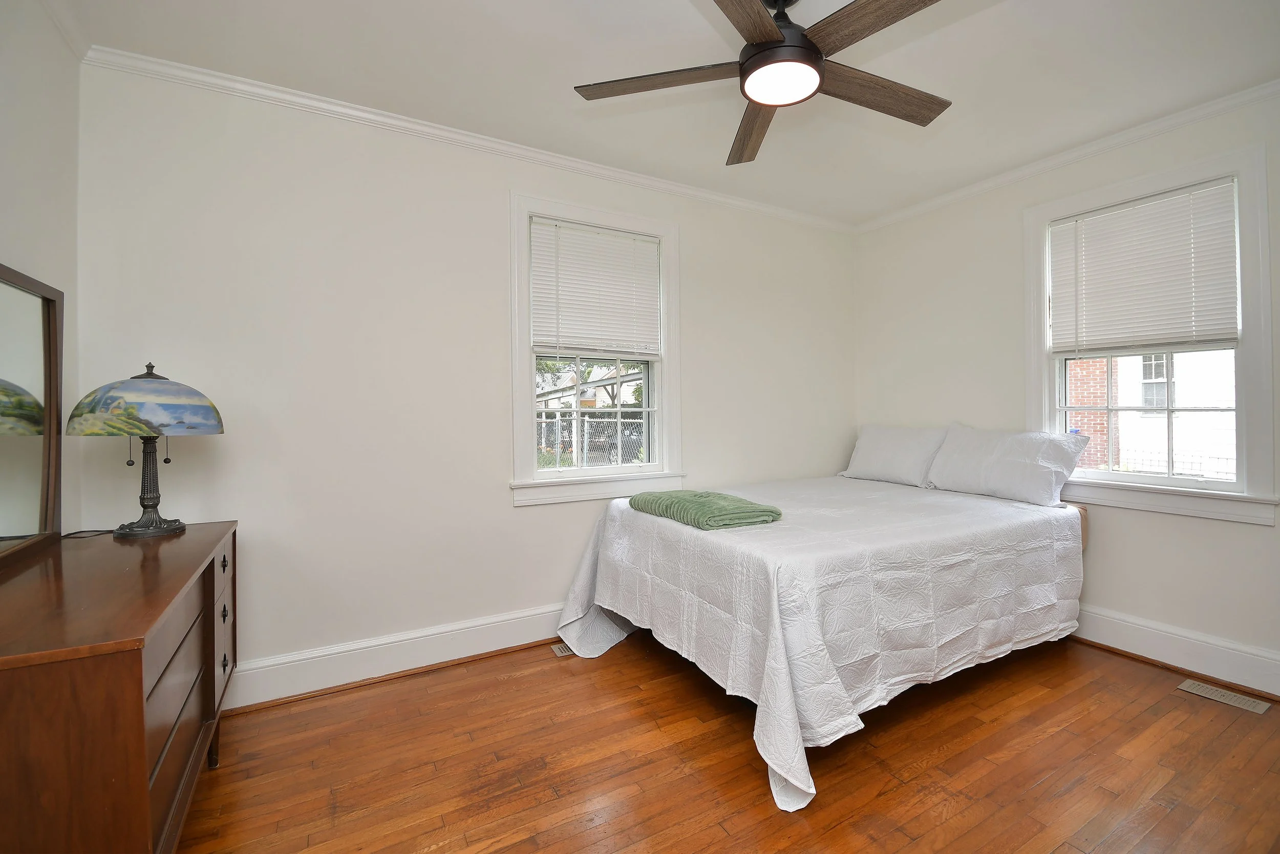 A bedroom with white walls, two windows with white blinds, a white bed with a white bedspread and pillows, a green blanket folded at the foot, a wooden dresser with a decorative lamp, a ceiling fan with wooden blades, and hardwood flooring.