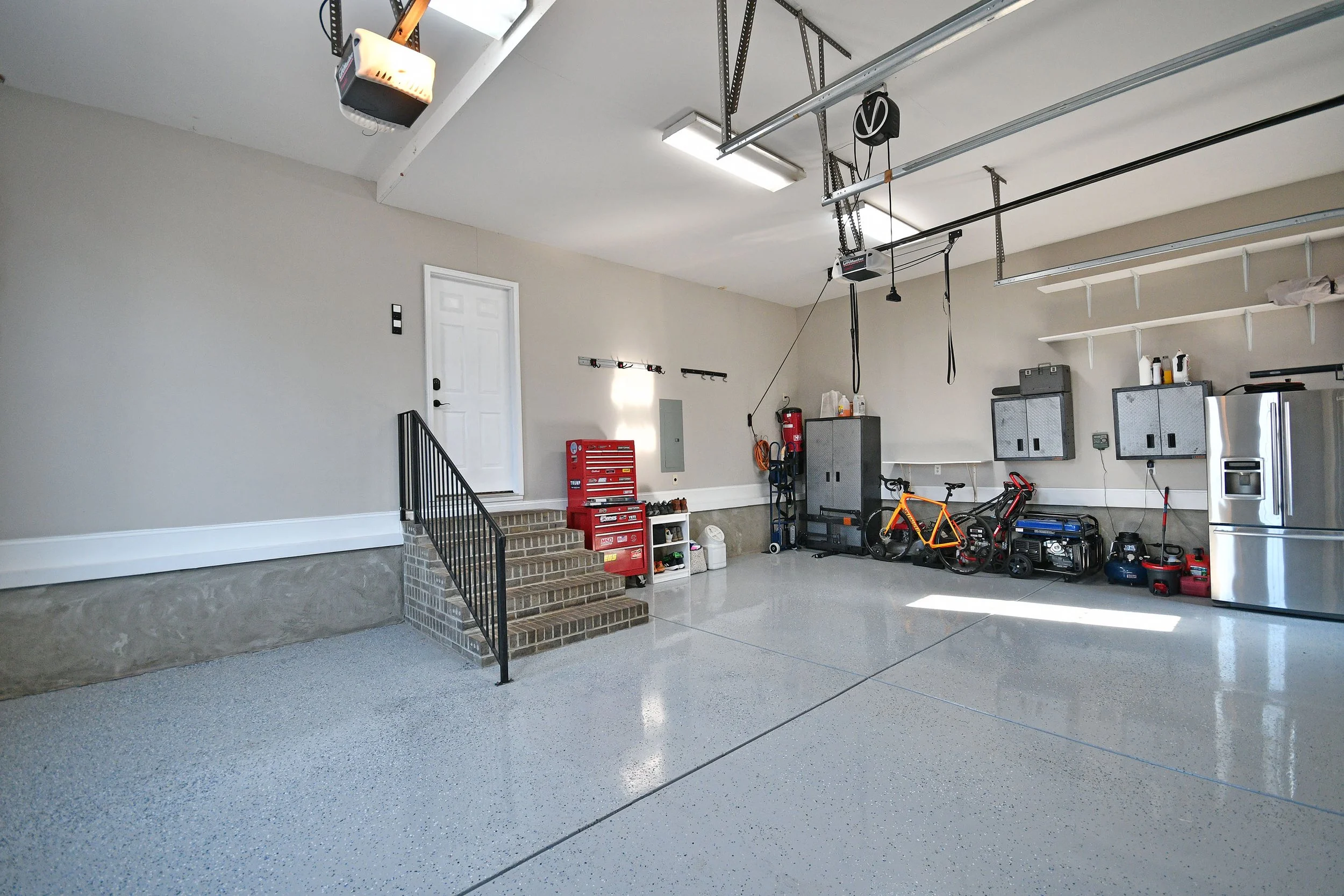 A clean, organized two-car garage with workout and storage areas, including bicycles, a red tool chest, cabinets, a refrigerator, and exercise equipment. Home for sale in Greenville, SC. Master on Main Level. 4 Bedroom home for sale. Upscale